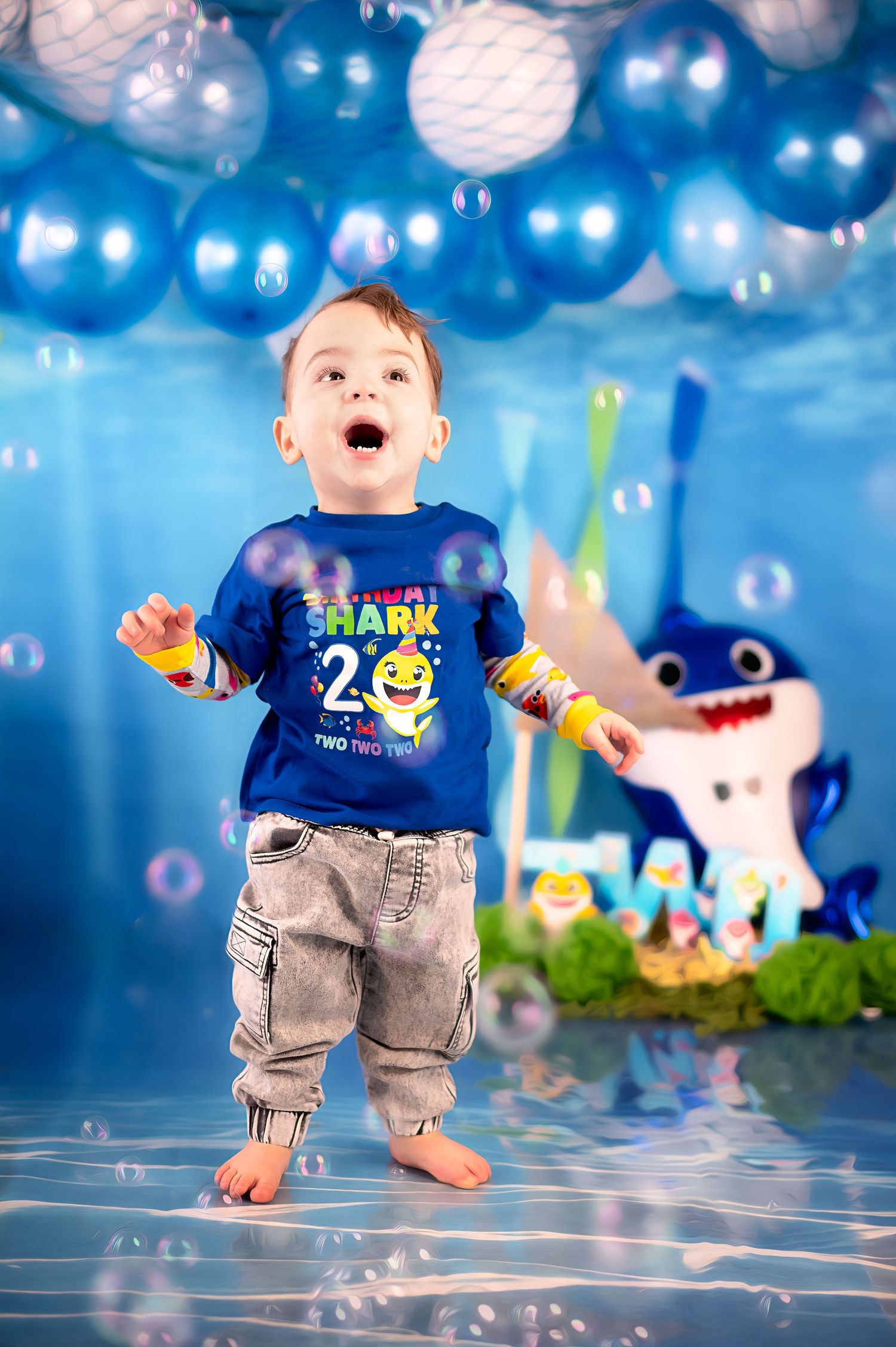 Toddler in shark shirt playing with bubbles against blue balloon backdrop.