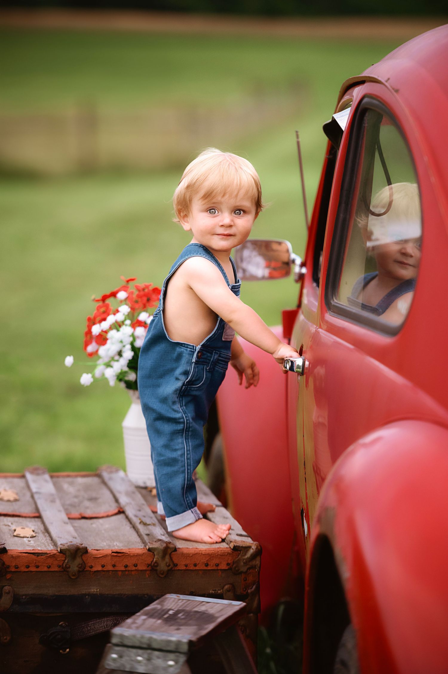 Young child in overalls climbing on wooden platform next to vintage red car.