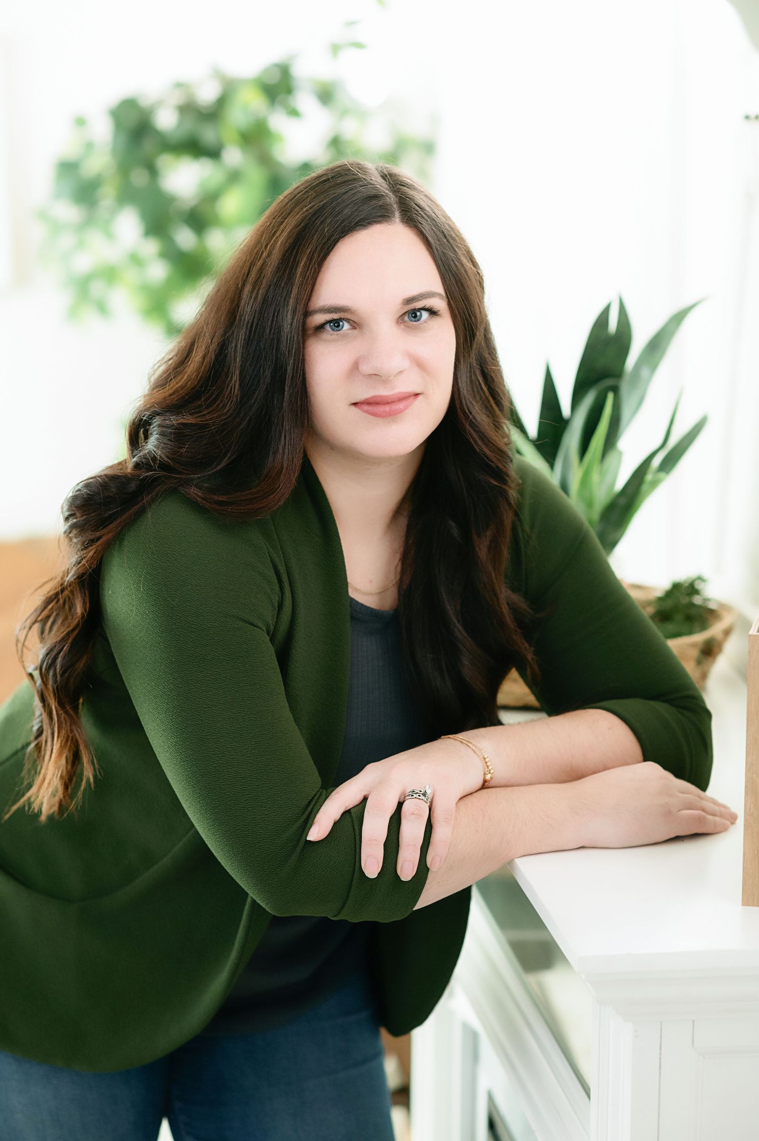 Portrait of a person in a dark green blazer leaning against a white surface with plants in the background.