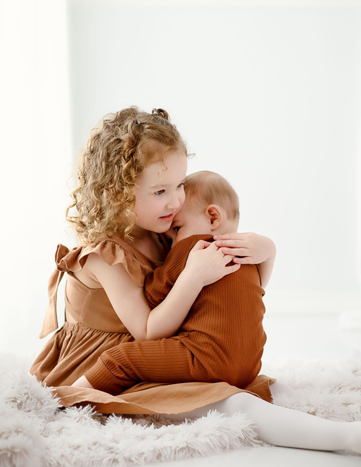 Two children in brown outfits share tender moments on white fluffy blanket.