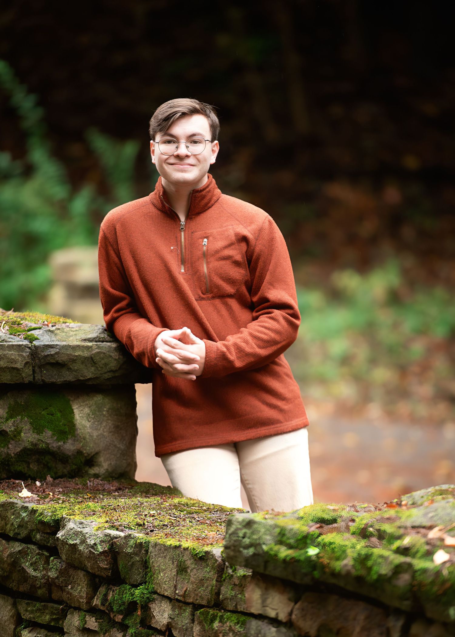 Person in rust-colored fleece sitting on mossy stone wall in autumn setting.