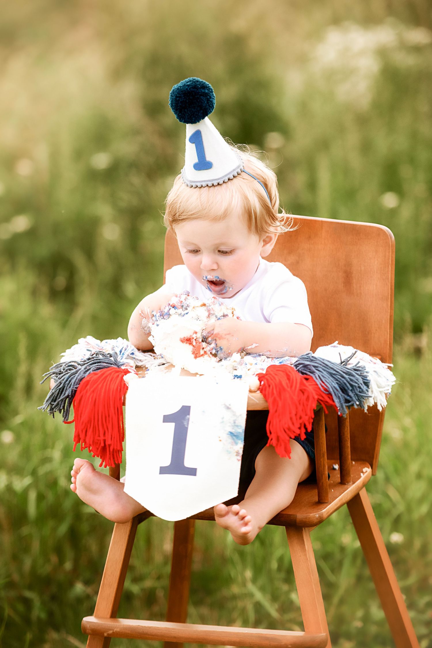 Baby wearing party hat enjoying cake smash during outdoor first birthday photoshoot.