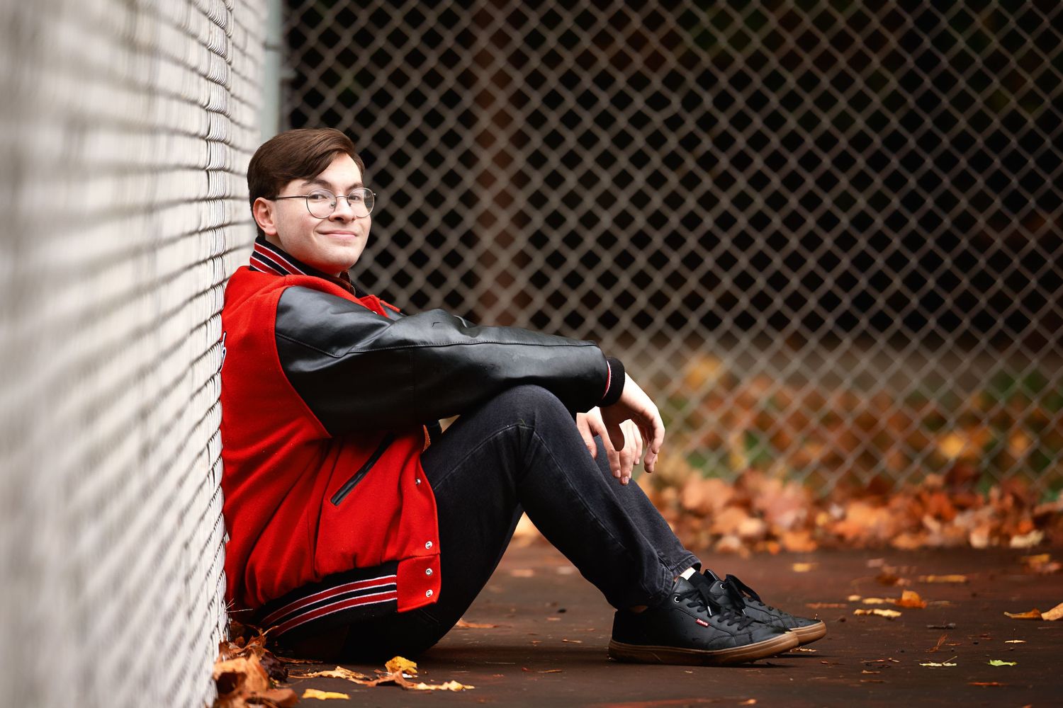 Person in red jacket relaxing against chain link fence with autumn leaves.