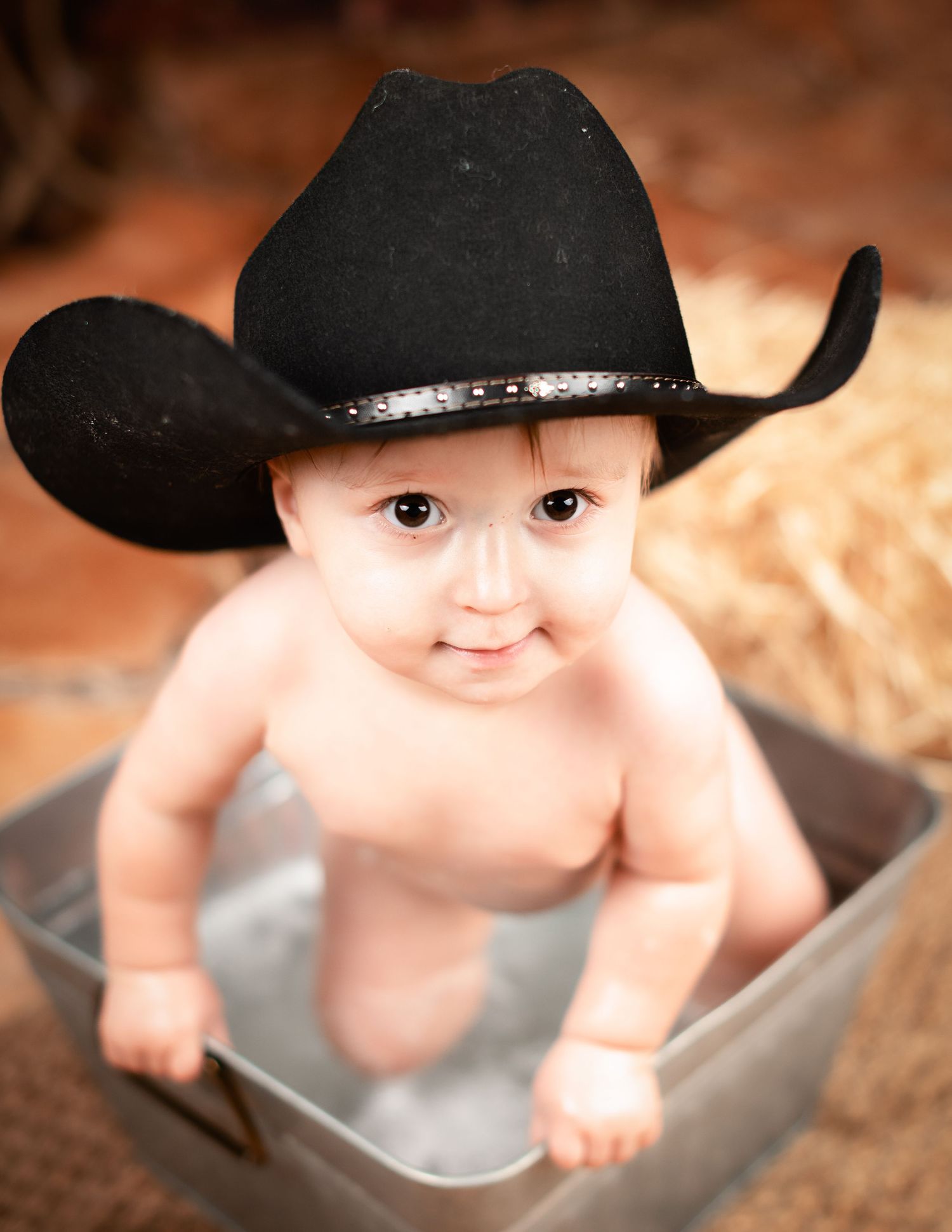 Adorable baby wearing a black cowboy hat sits in a metal bucket against a warm brown background.