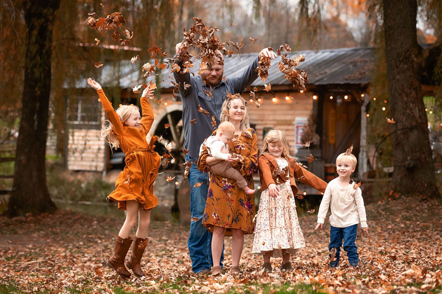 Autumn family photography session with falling leaves and glowing string lights in a wooded setting.