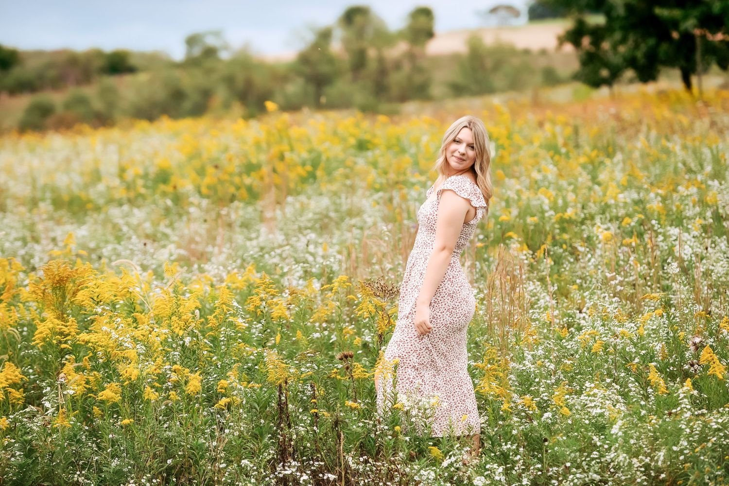 Woman in flowing pink dress twirls in a field of yellow wildflowers on a sunny day.