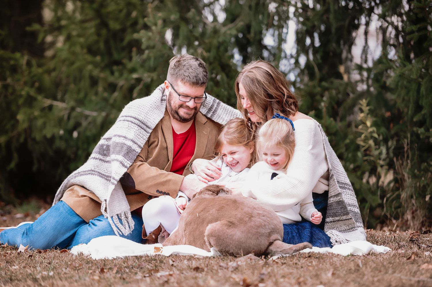 A couple and their dog lay together on a blanket in a natural outdoor setting during a family photo session.
