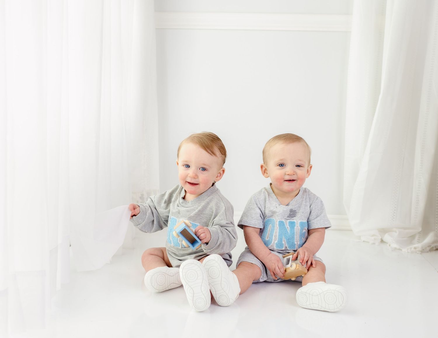 Two babies in matching grey outfits sitting against white curtain background.