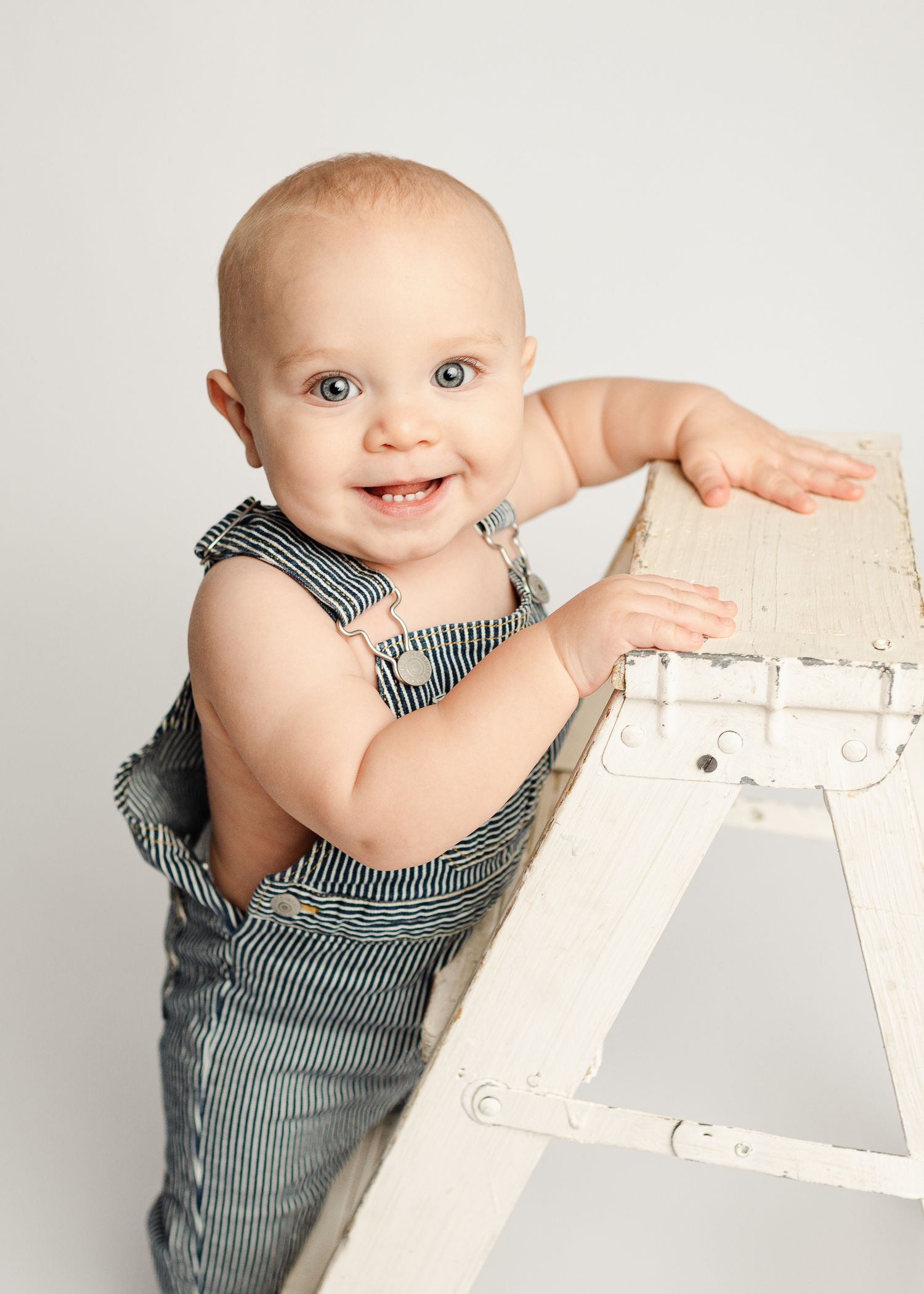 A smiling baby in striped overalls holds onto a white wooden chair while crawling and playing.