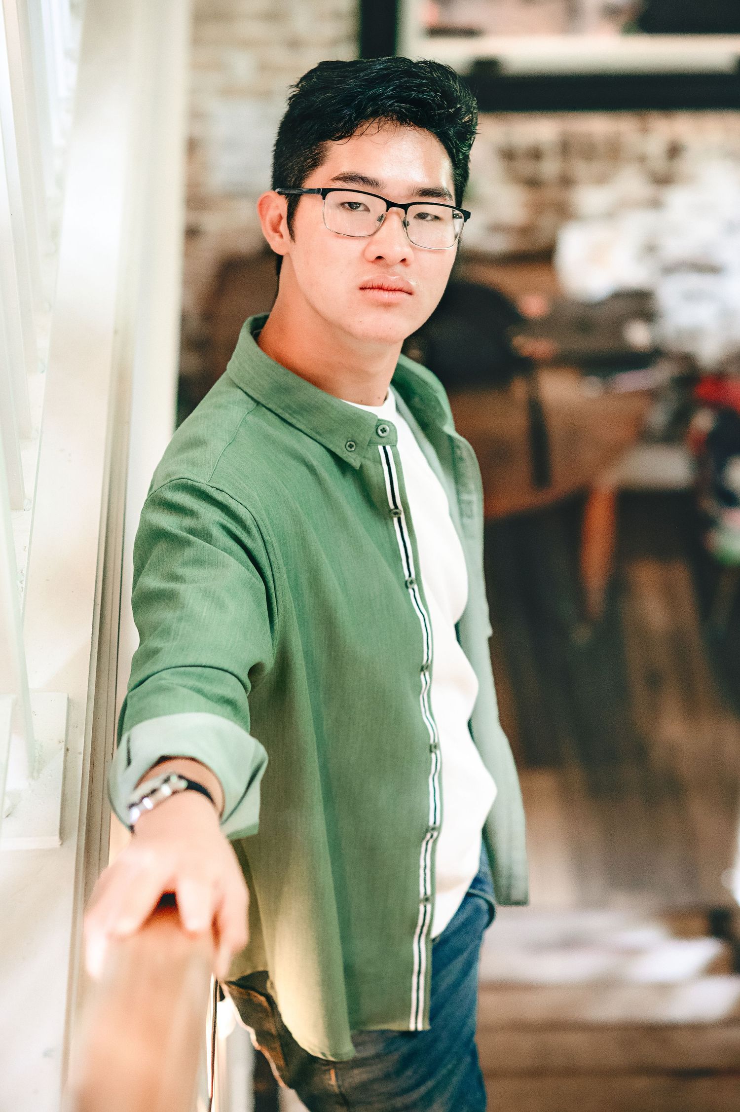 Student in green button-down shirt and glasses leans against wall in casual indoor setting.