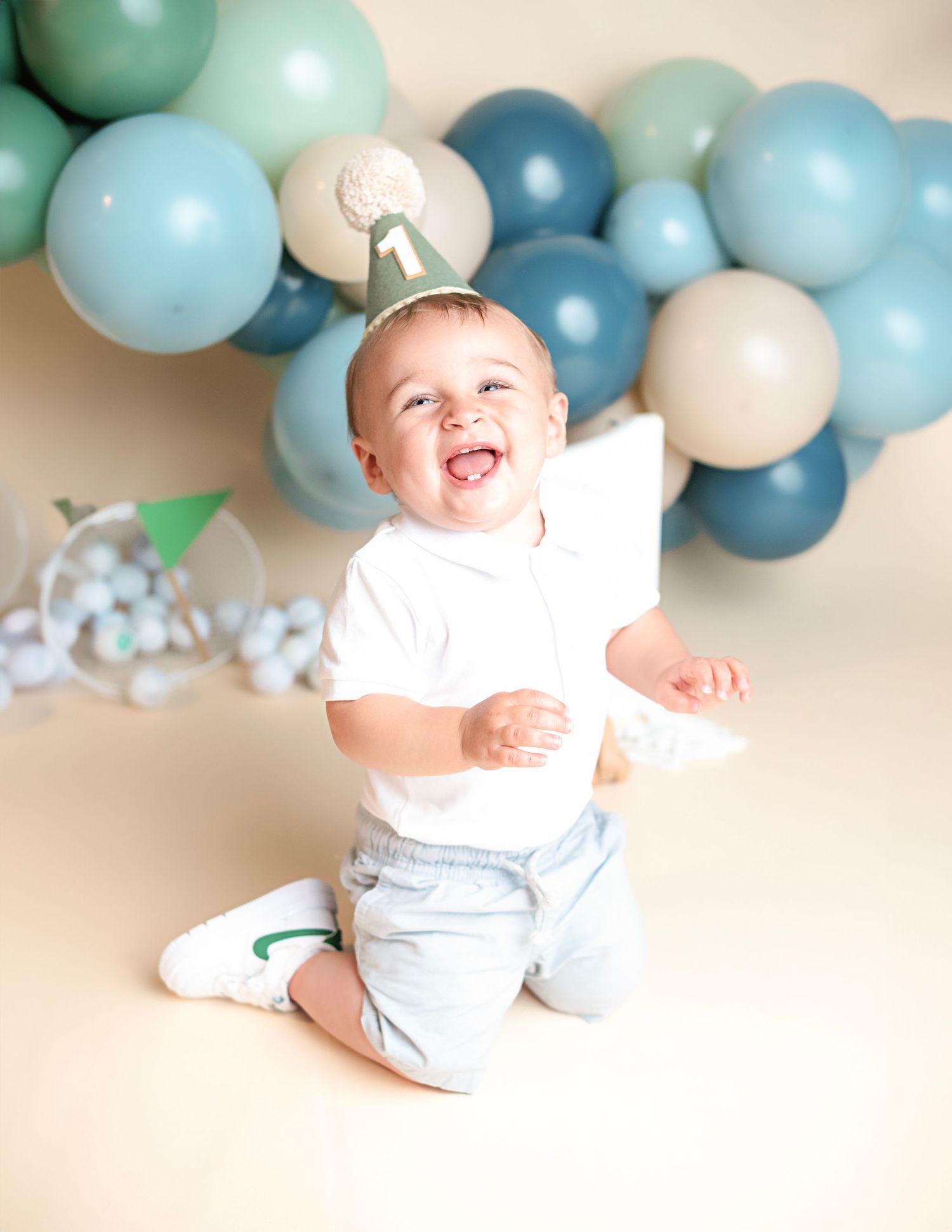 Baby in white outfit sits among soft blue and mint green balloons at a festive celebration.