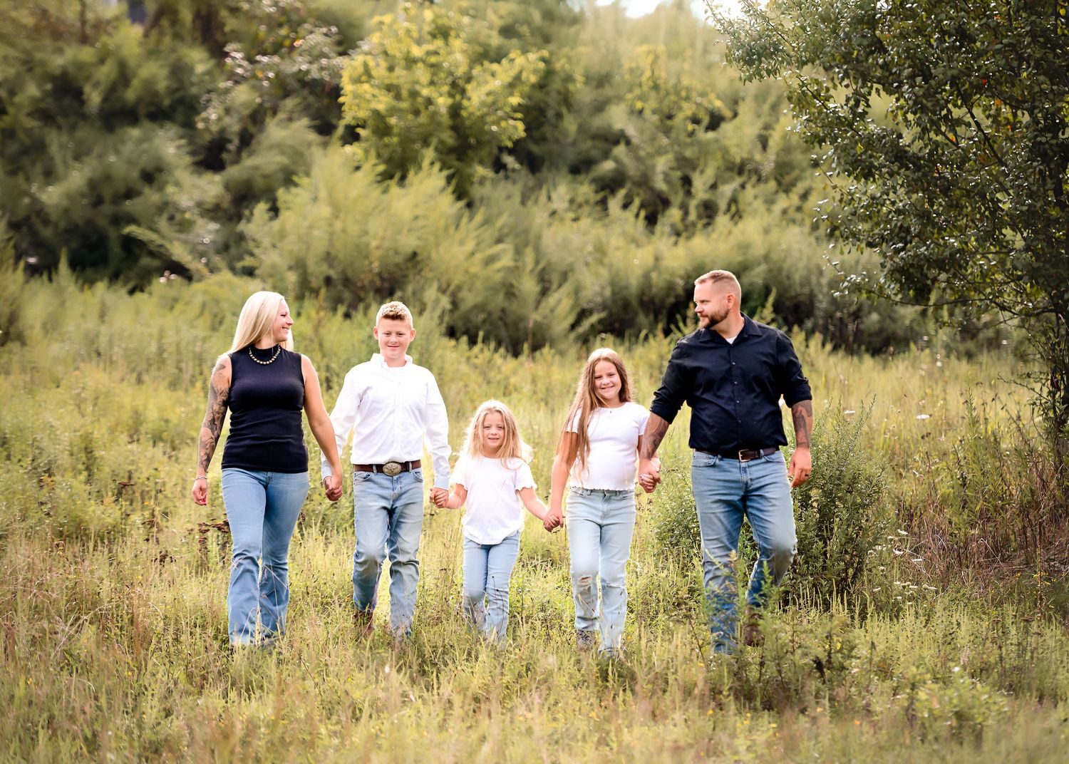 Family walking together through meadow holding hands in casual denim outfits.
