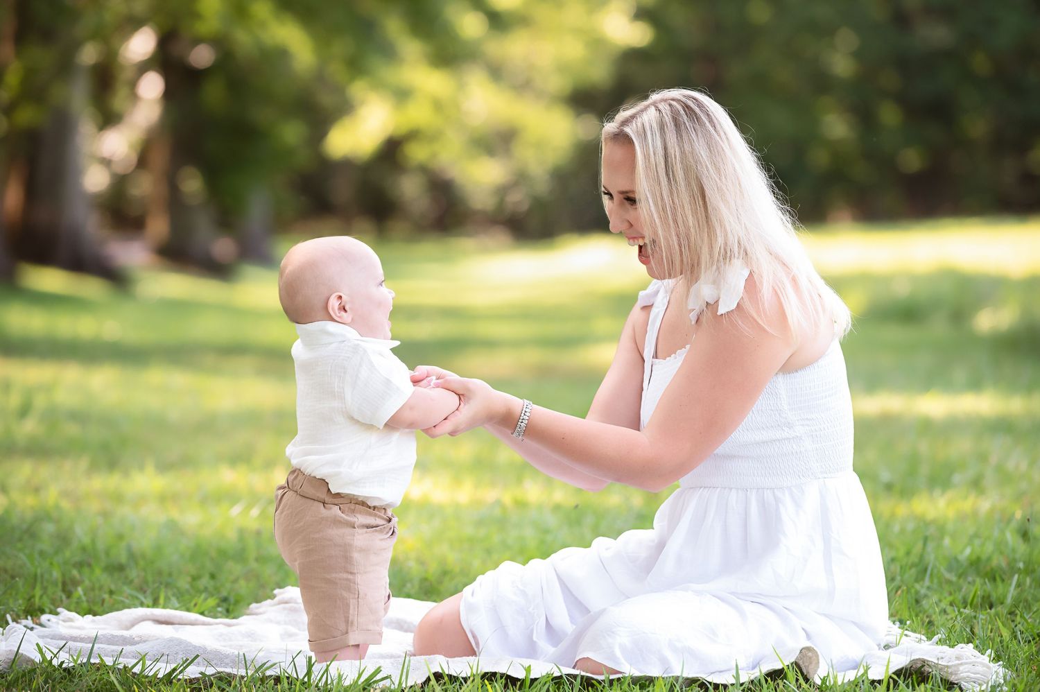 Mother and baby in white outfits playing together on sunny grass outdoors.