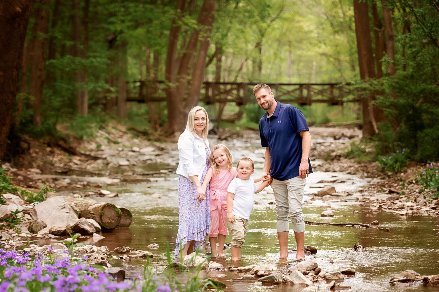 A family stands together in a scenic creek bed surrounded by lush forest and purple wildflowers on a summer day.