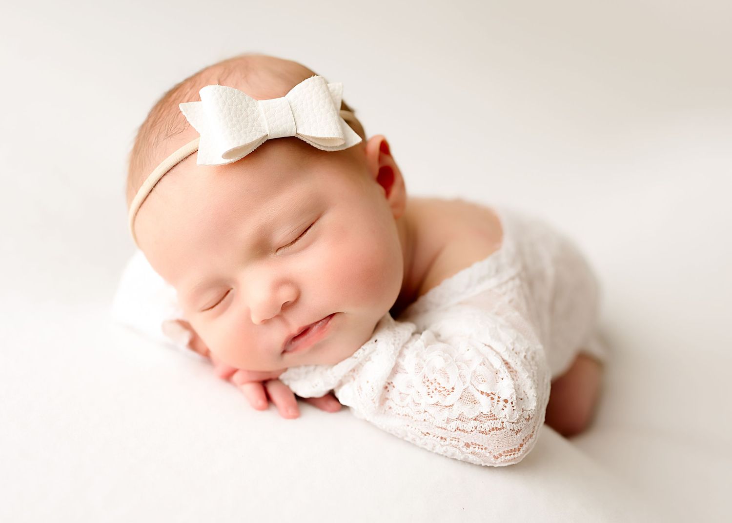 A sleeping newborn baby in white lace swaddle with bow headband photographed in a soft light studio setting.