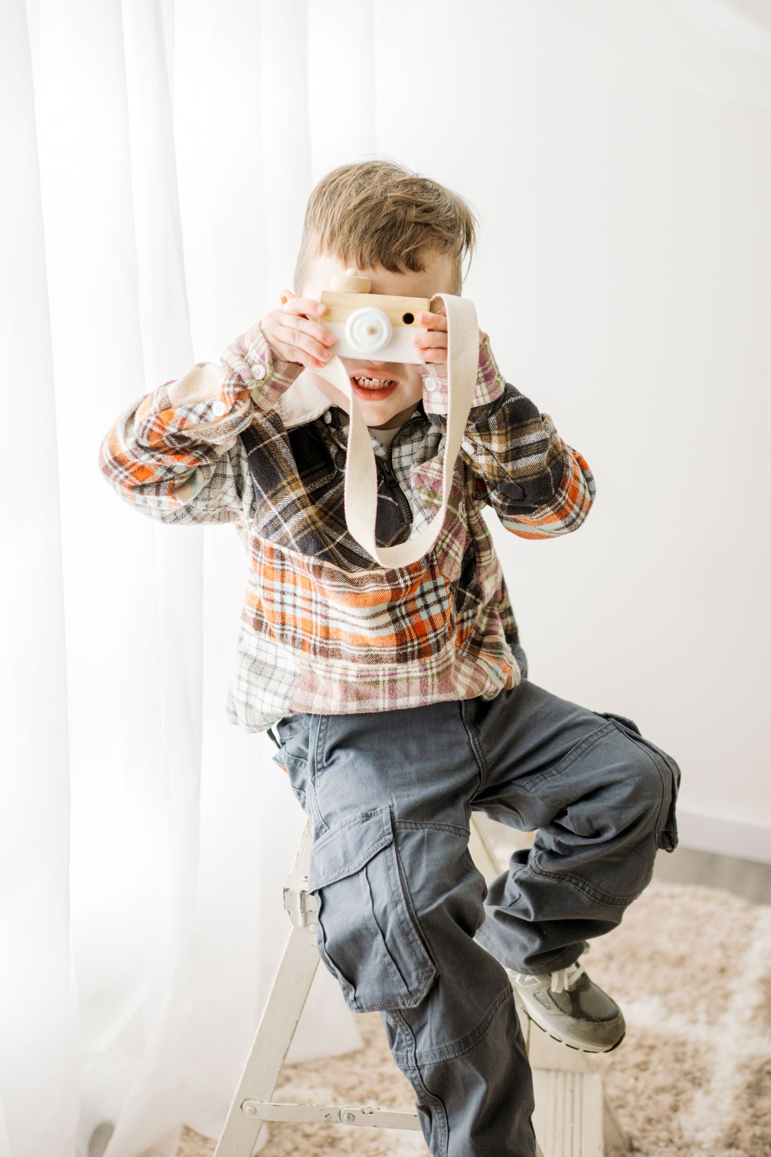 Young child in plaid shirt and jeans plays with white toilet paper roll while sitting on carpeted floor.
