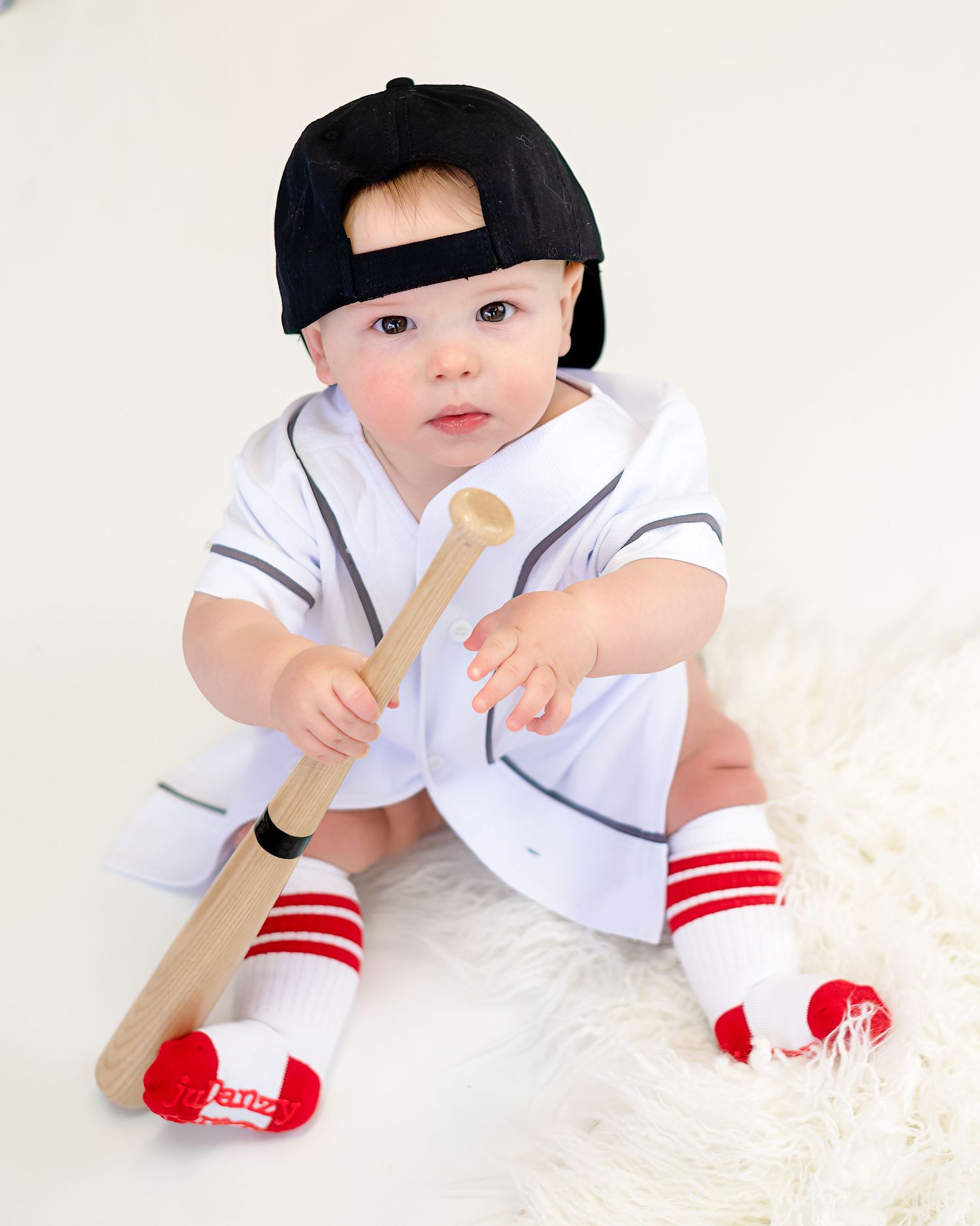 Baby in baseball uniform with black cap holding wooden bat on white background.