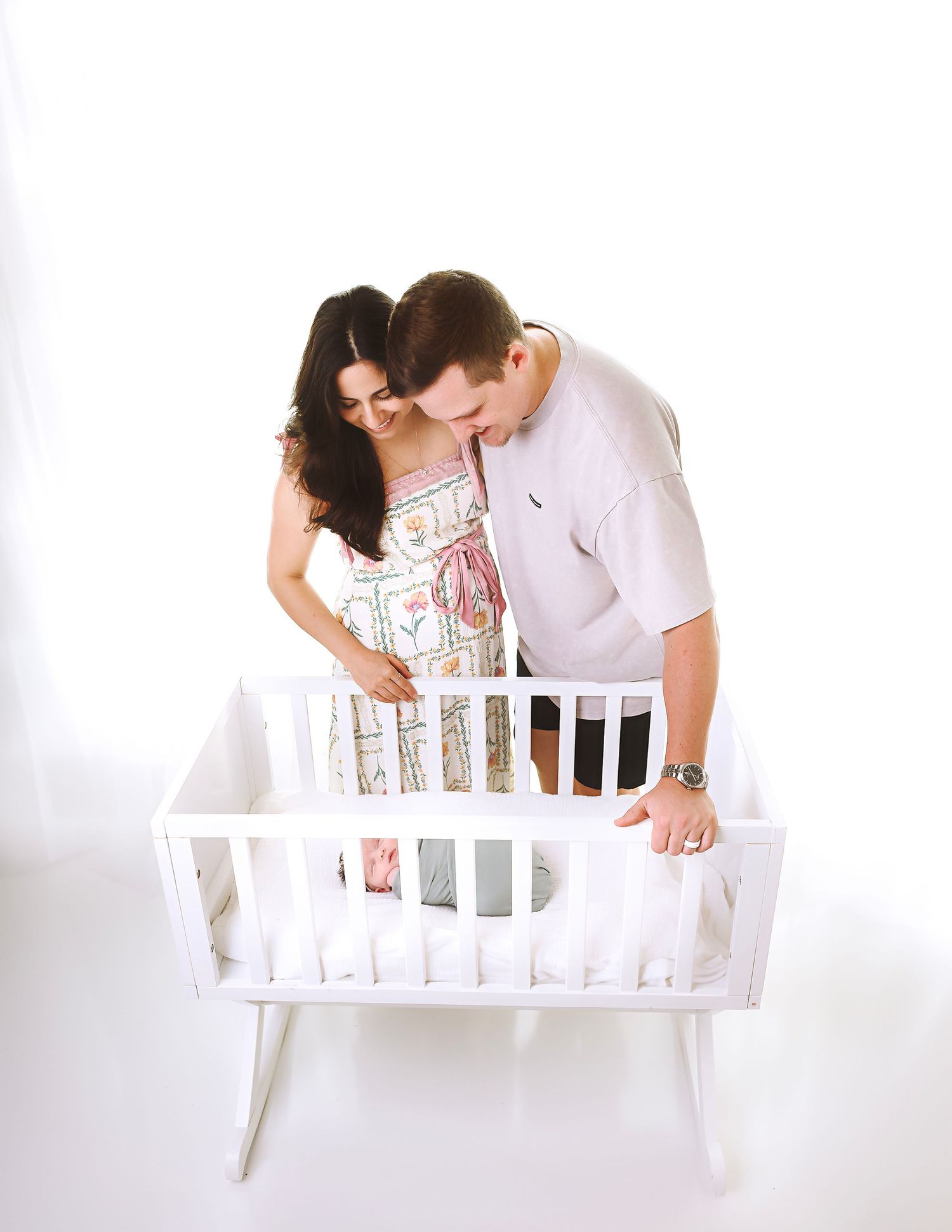 Parents looking lovingly at baby in white crib against white background.