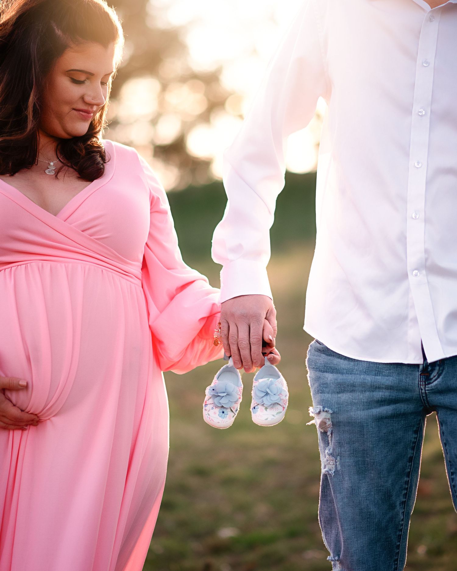 Couple holding baby shoes during sunset, woman wearing flowing pink maternity dress.