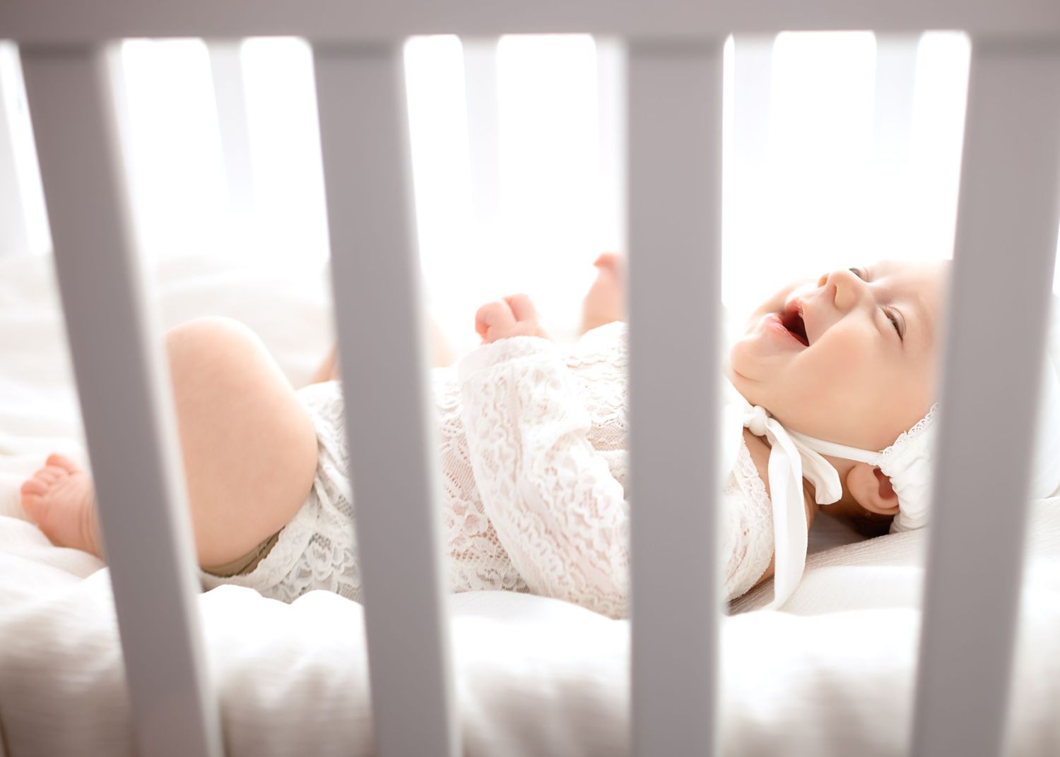 View through crib slats of baby resting on white bedding in natural light.