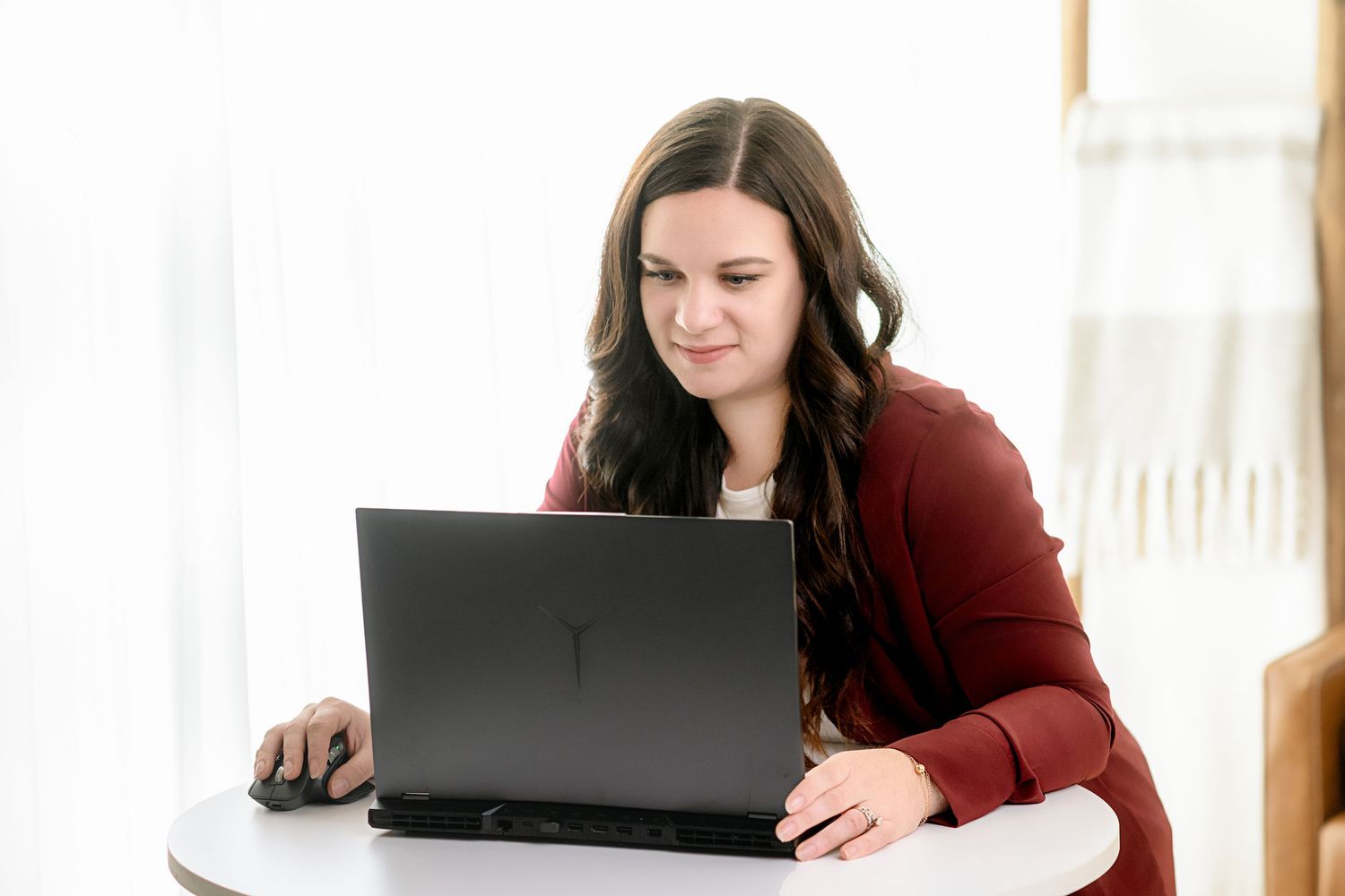Woman in burgundy sweater working on laptop in bright sunlit room.
