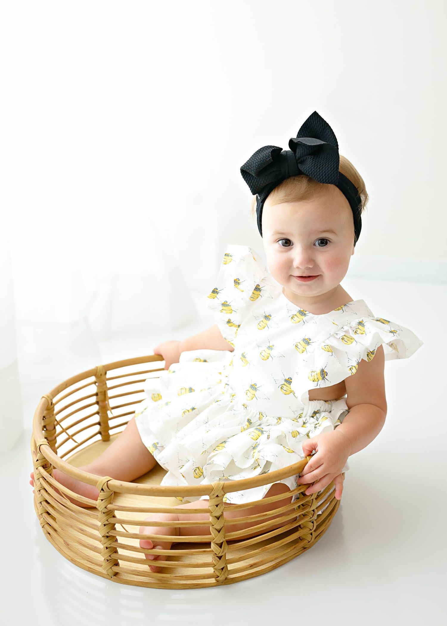 Baby wearing floral dress and black bow sits in wooden basket against white backdrop.