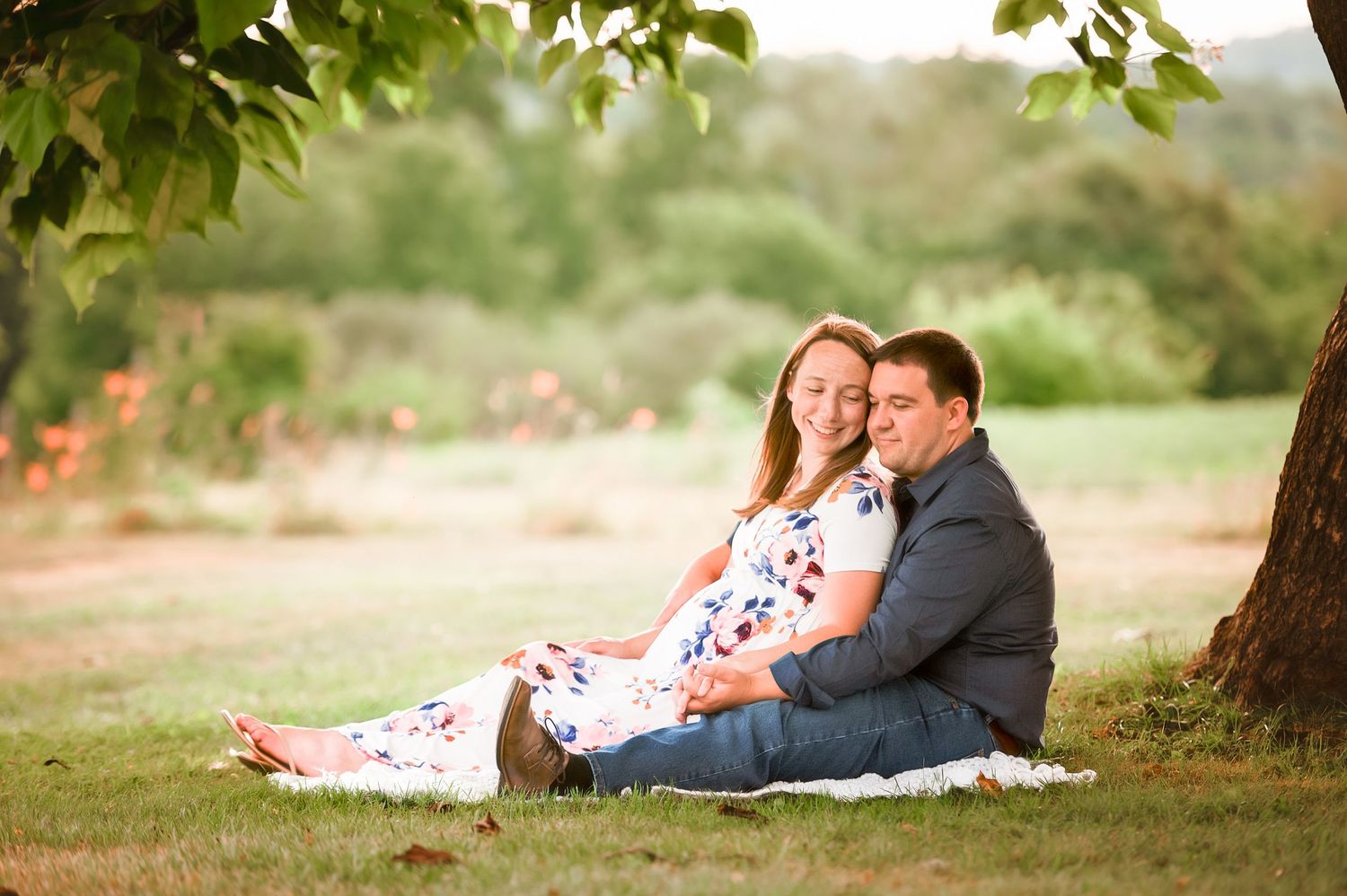 A couple relaxes together on a blanket under a shady tree in a park setting.