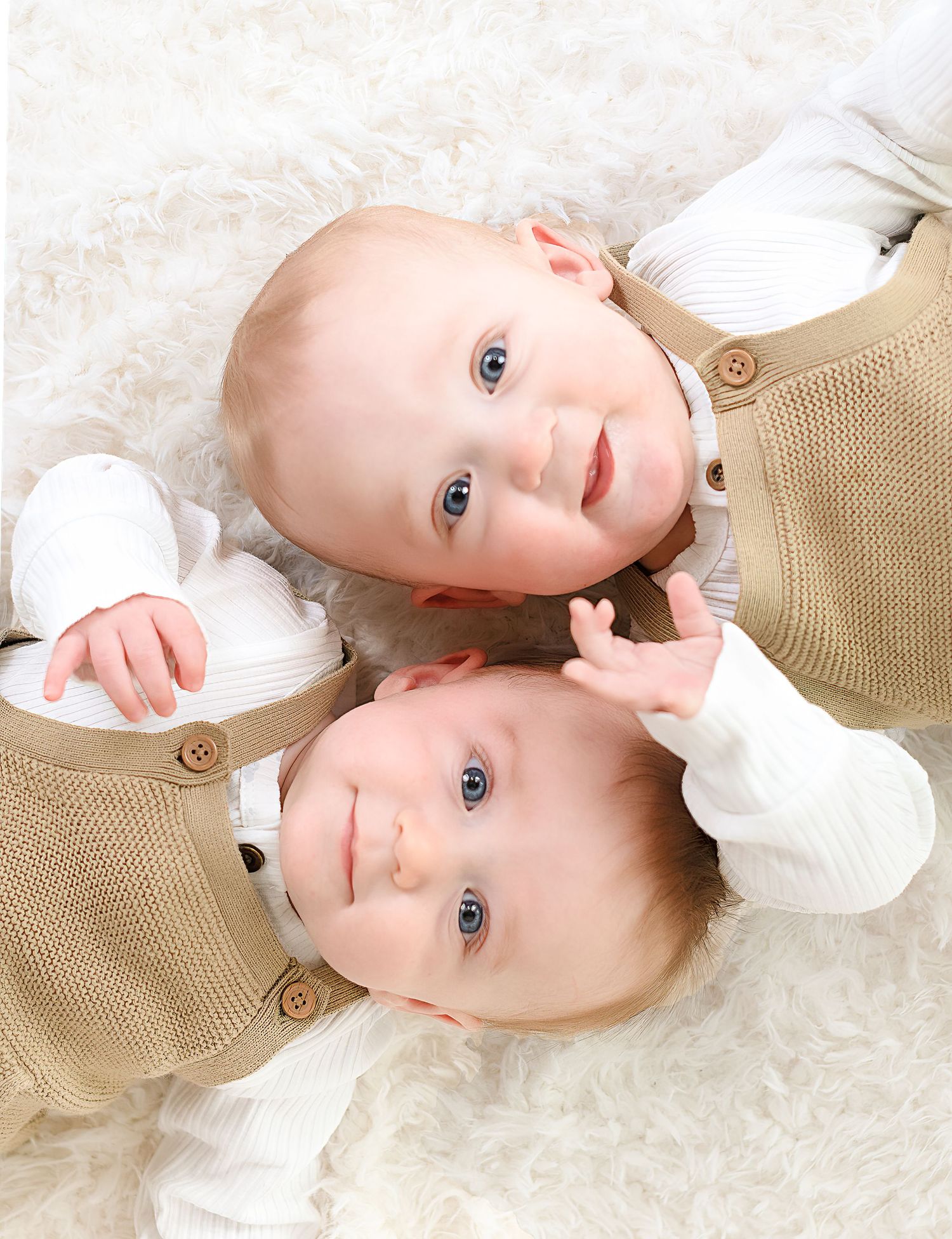 Two babies in white outfits lying on cream textured background.