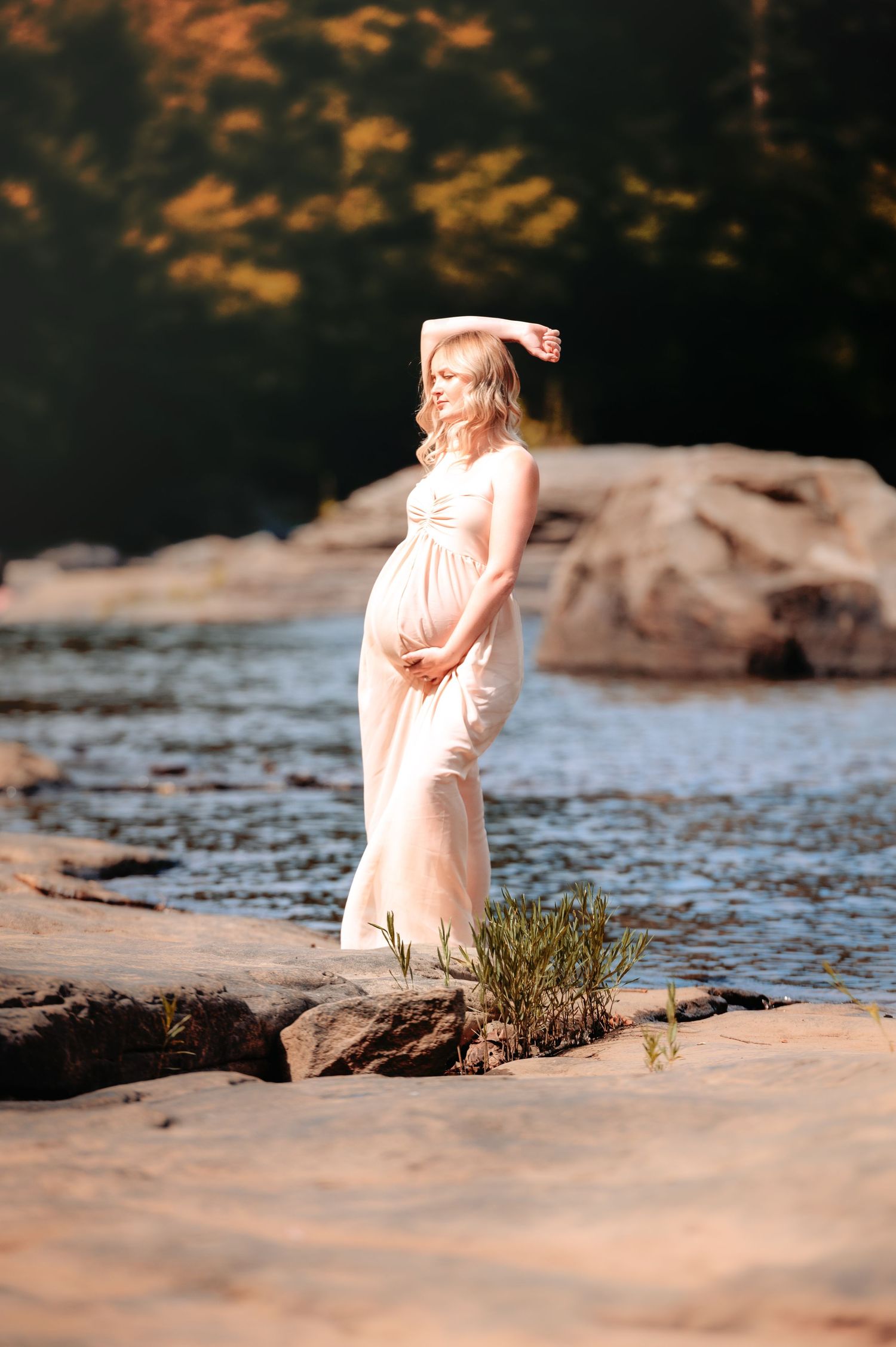 Peaceful riverside maternity portrait with soft lighting and rocky landscape.