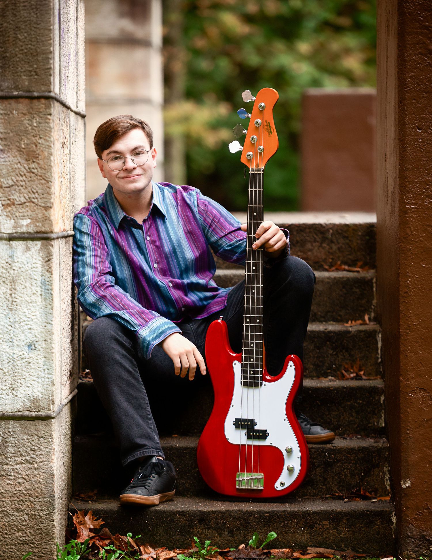 Musician posing with red bass guitar on stone steps surrounded by fall foliage.