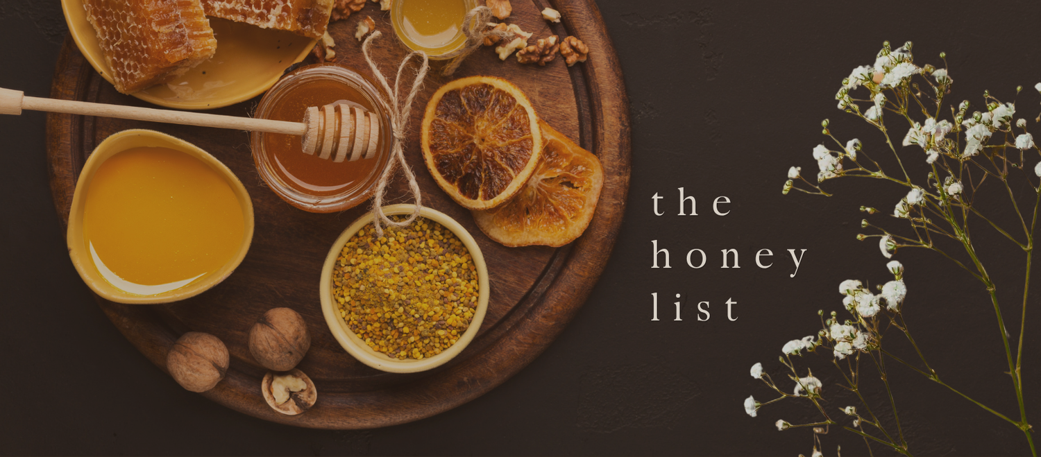 A wooden board with honey, honeycomb, dried citrus, walnuts, and bee pollen beside white flowers on a dark background.