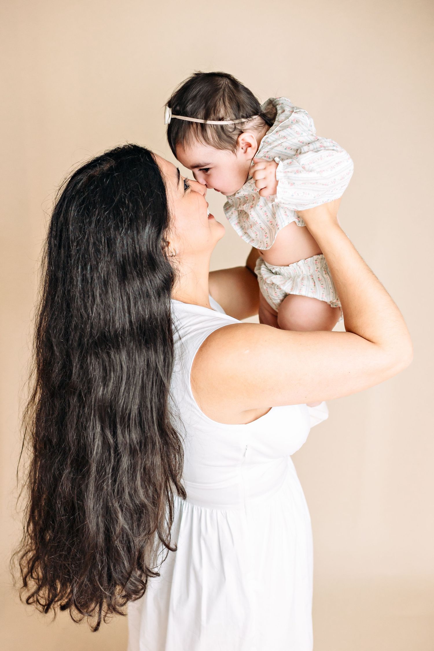 Mother and child in white dresses sharing tender moment against beige background.
