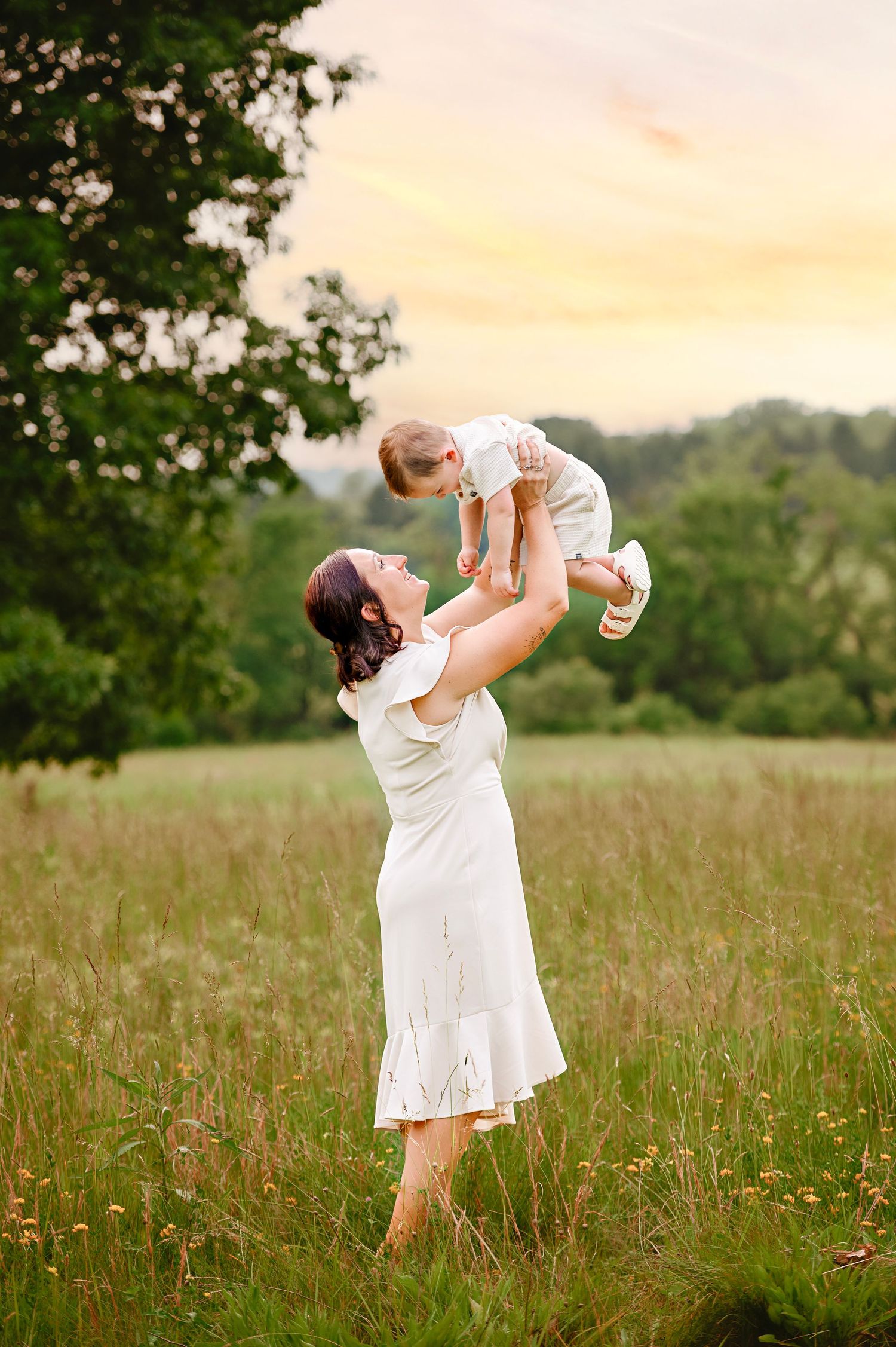 Mother lifting baby joyfully in wildflower field during golden sunset.