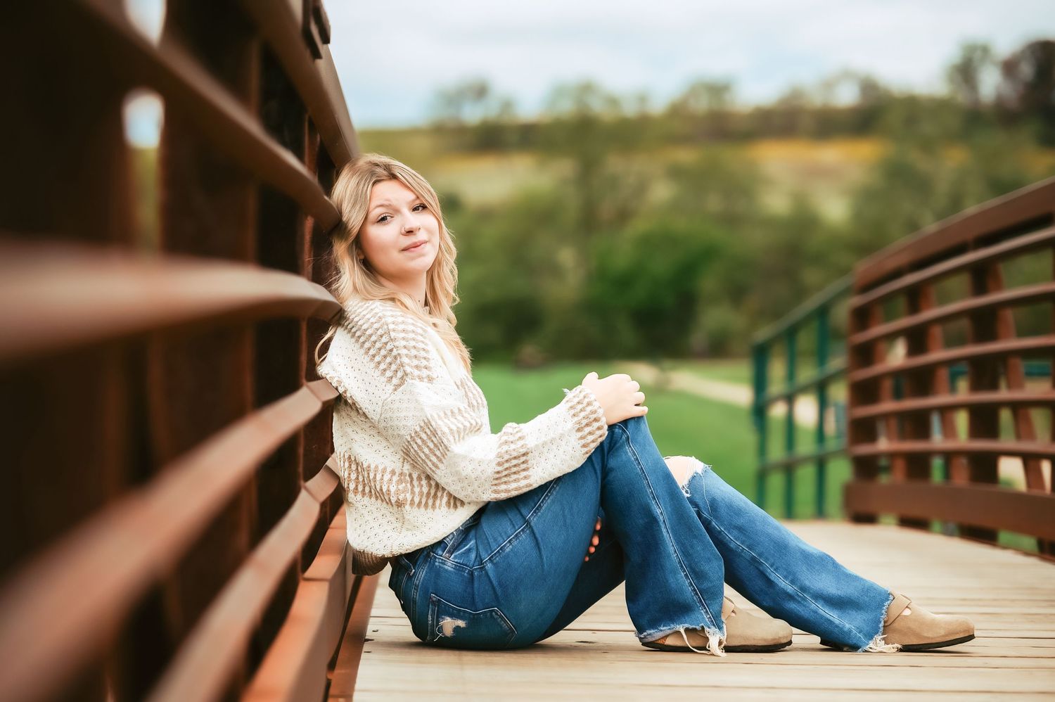 Relaxed pose against wooden bridge railing in cream sweater and jeans.