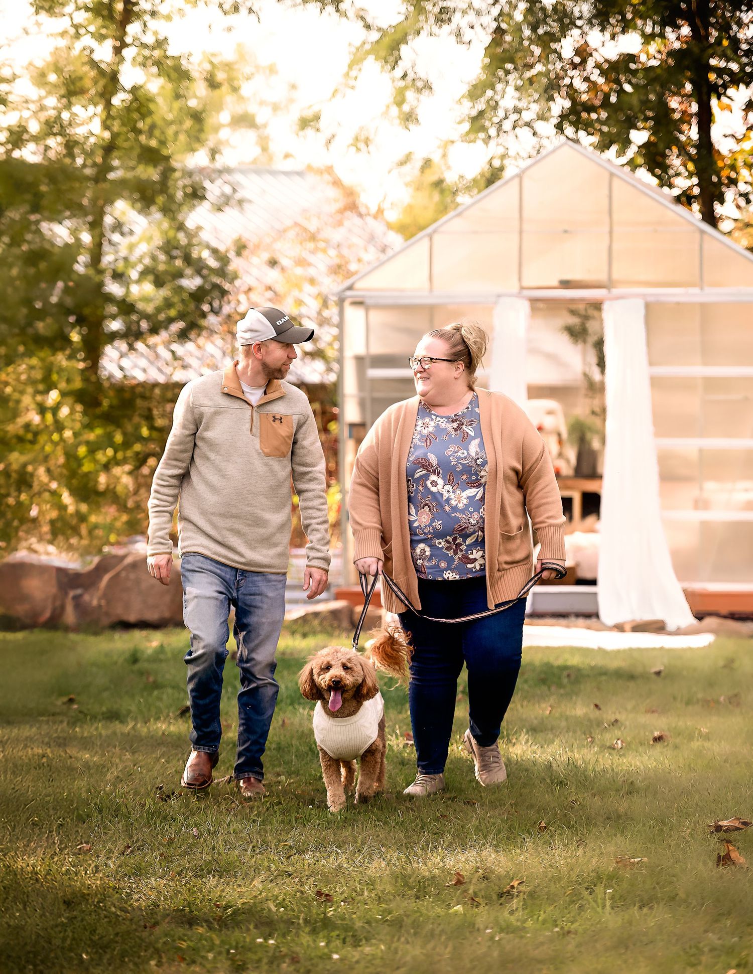 A couple walks their golden retriever dog across a grassy yard near a greenhouse.