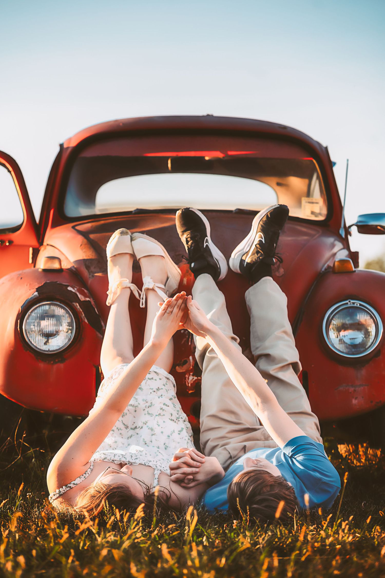 Playful couple poses upside down in vintage red car surrounded by wildflowers at sunset.