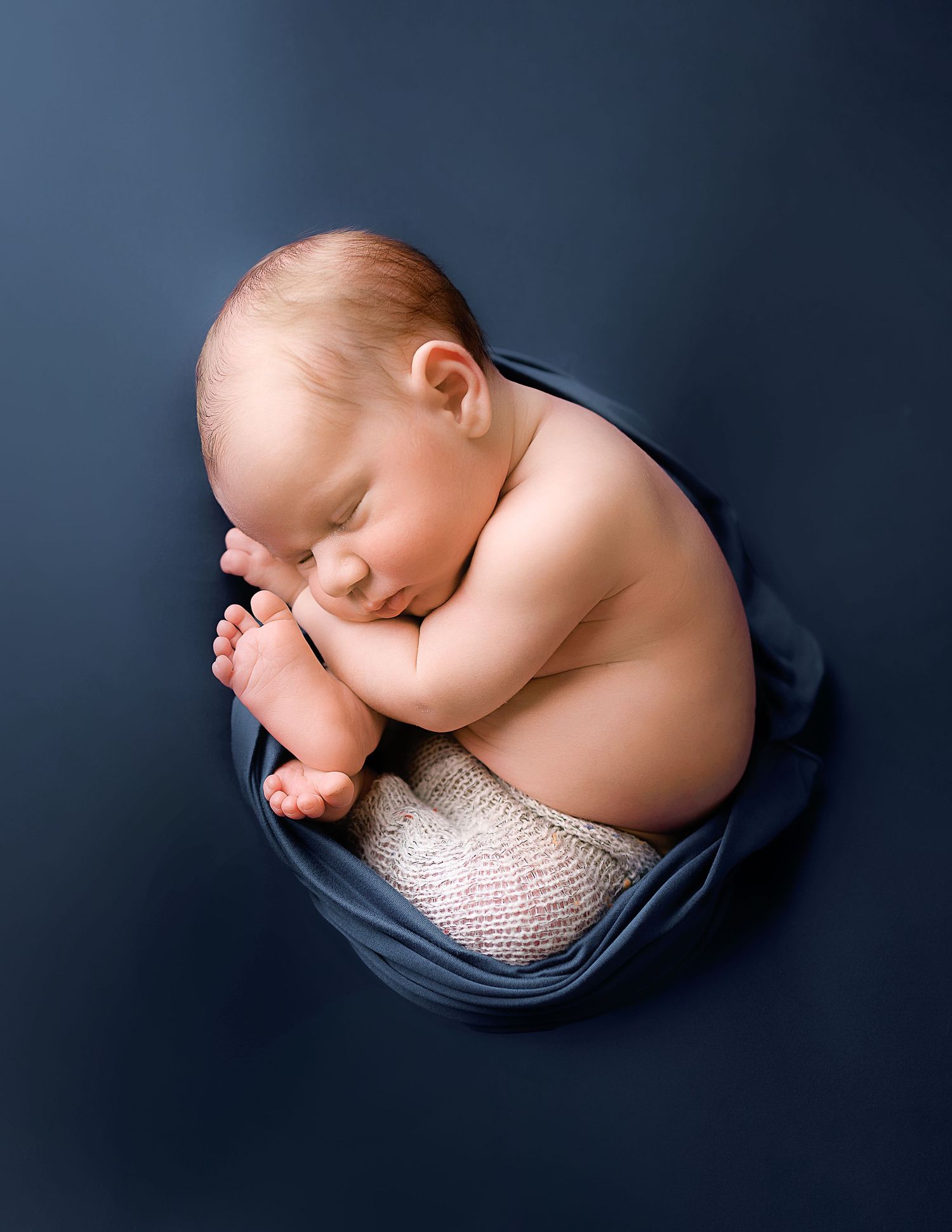 Sleeping newborn wrapped in navy blue fabric against dark background during professional photo session.
