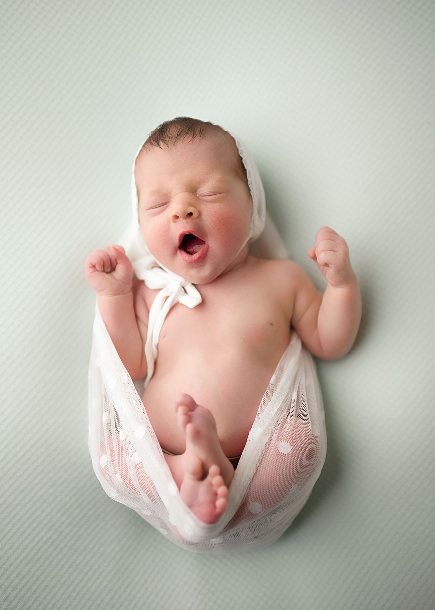 Newborn stretches and yawns while lying on soft mint green background.