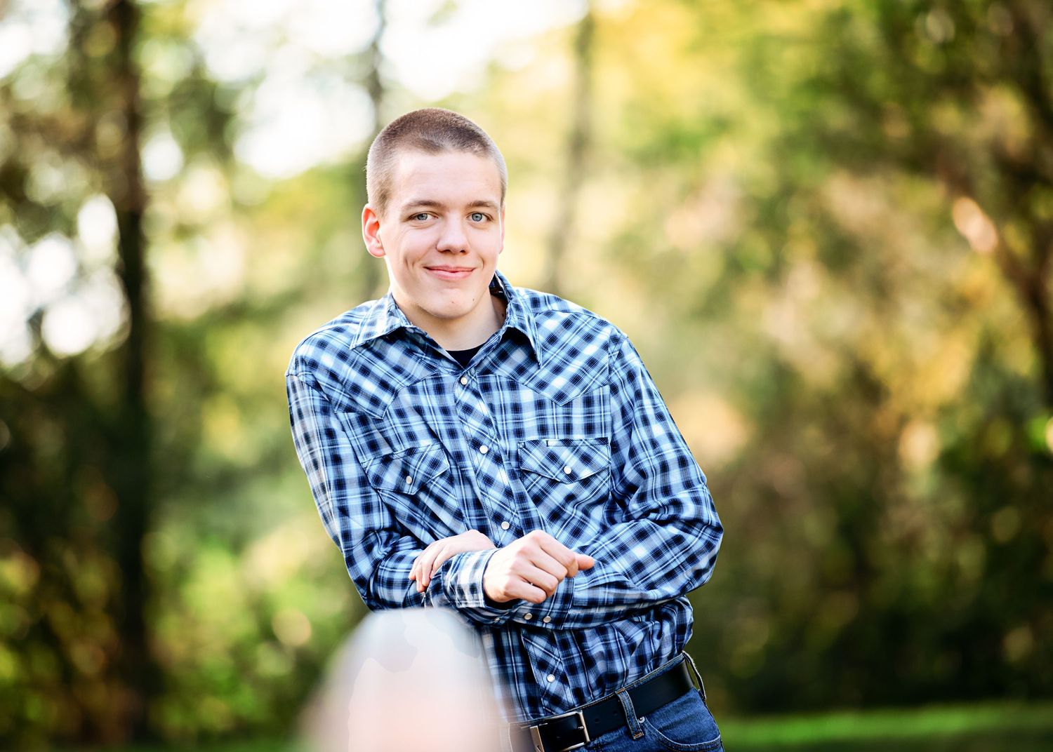 Outdoor portrait in blue plaid shirt with natural lighting and greenery background.