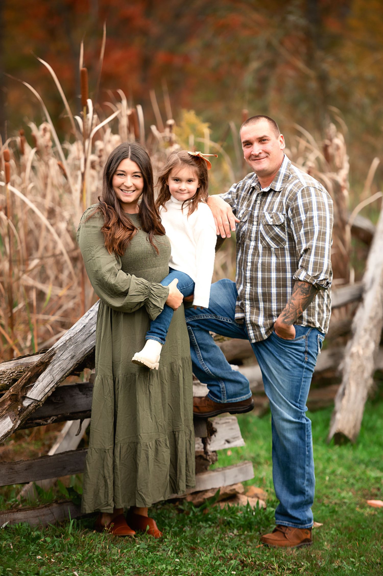 A family poses together by a rustic wooden fence with cattails and fall colors in the background.