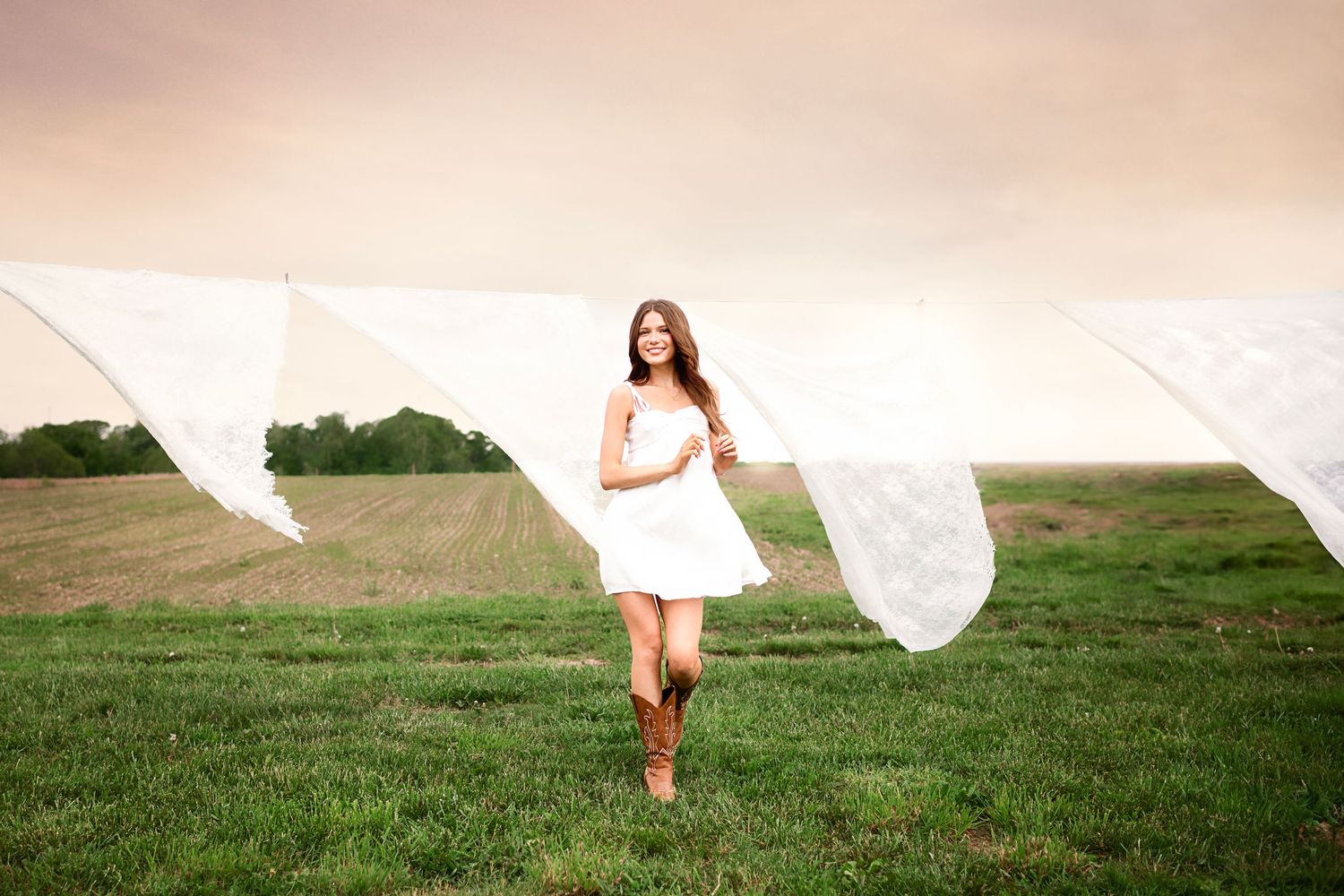 Woman in flowing white dress walking through green grass field at sunset.