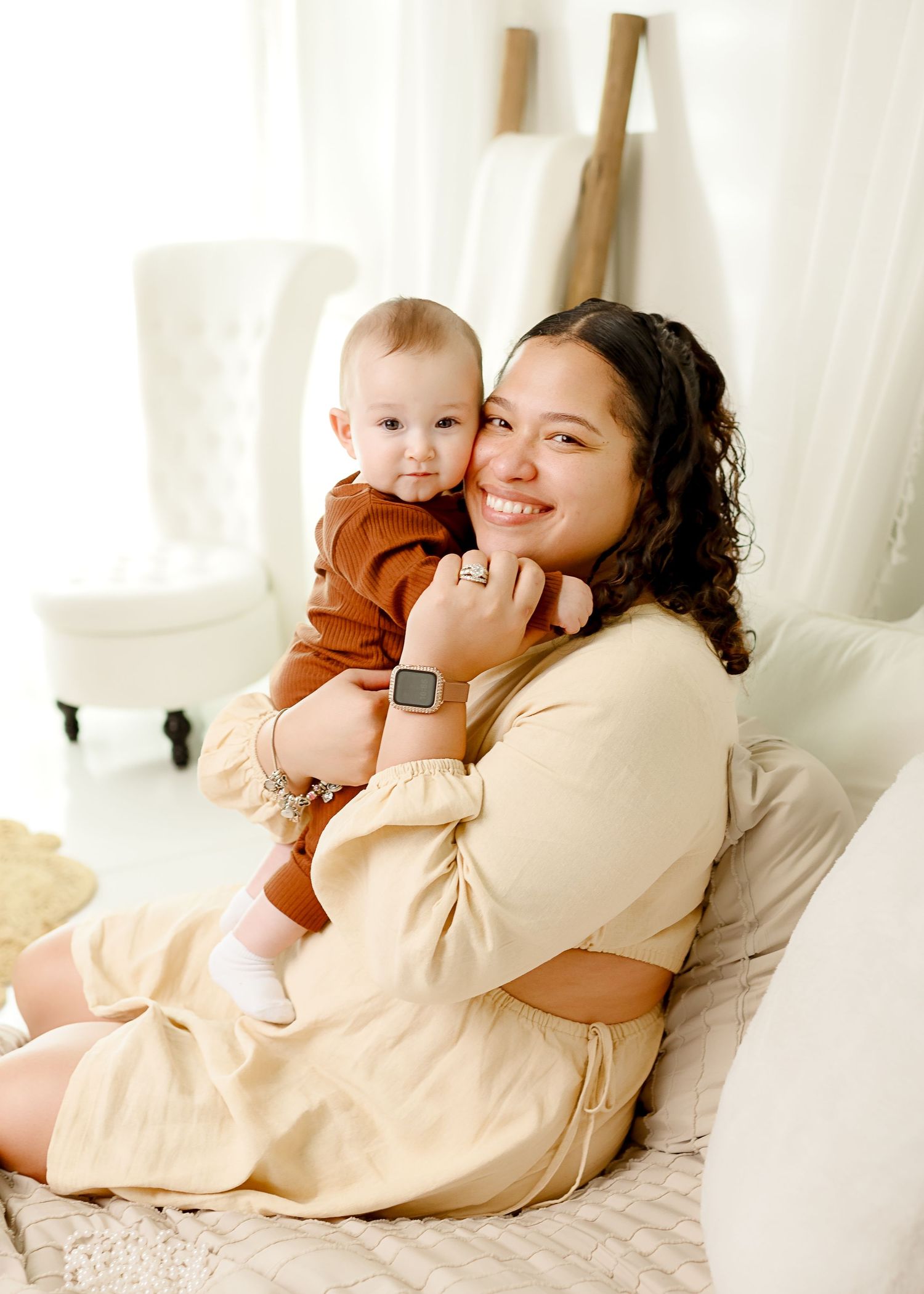 Parent and baby share a tender moment while sitting on white couch with decorative ladder in background.