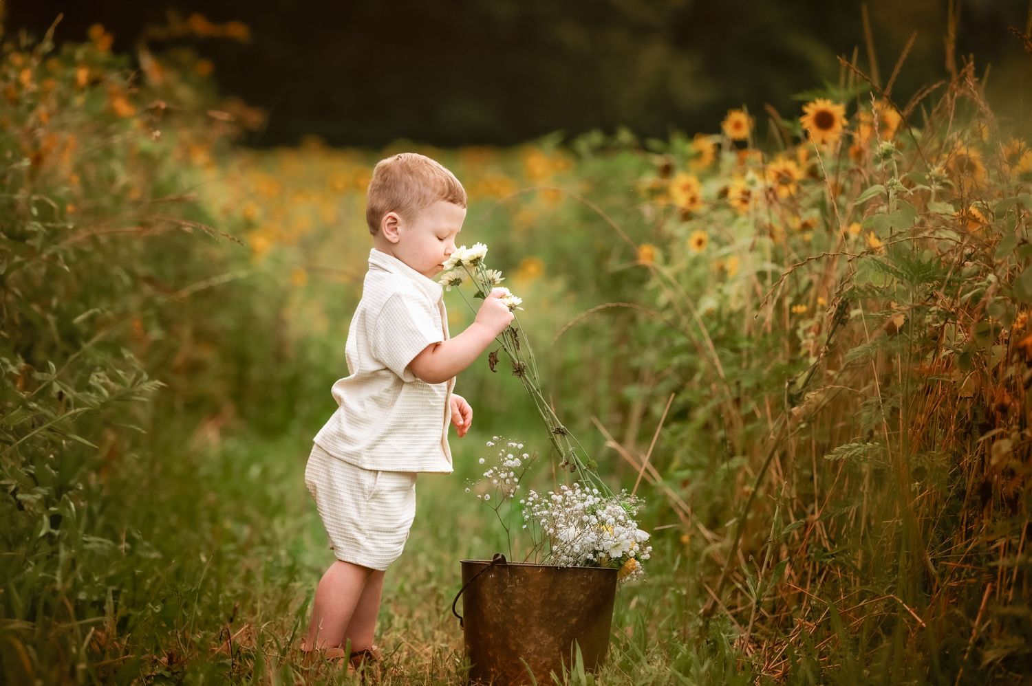 A child in white clothing explores a wildflower garden on a summer day with a rustic metal bucket nearby.