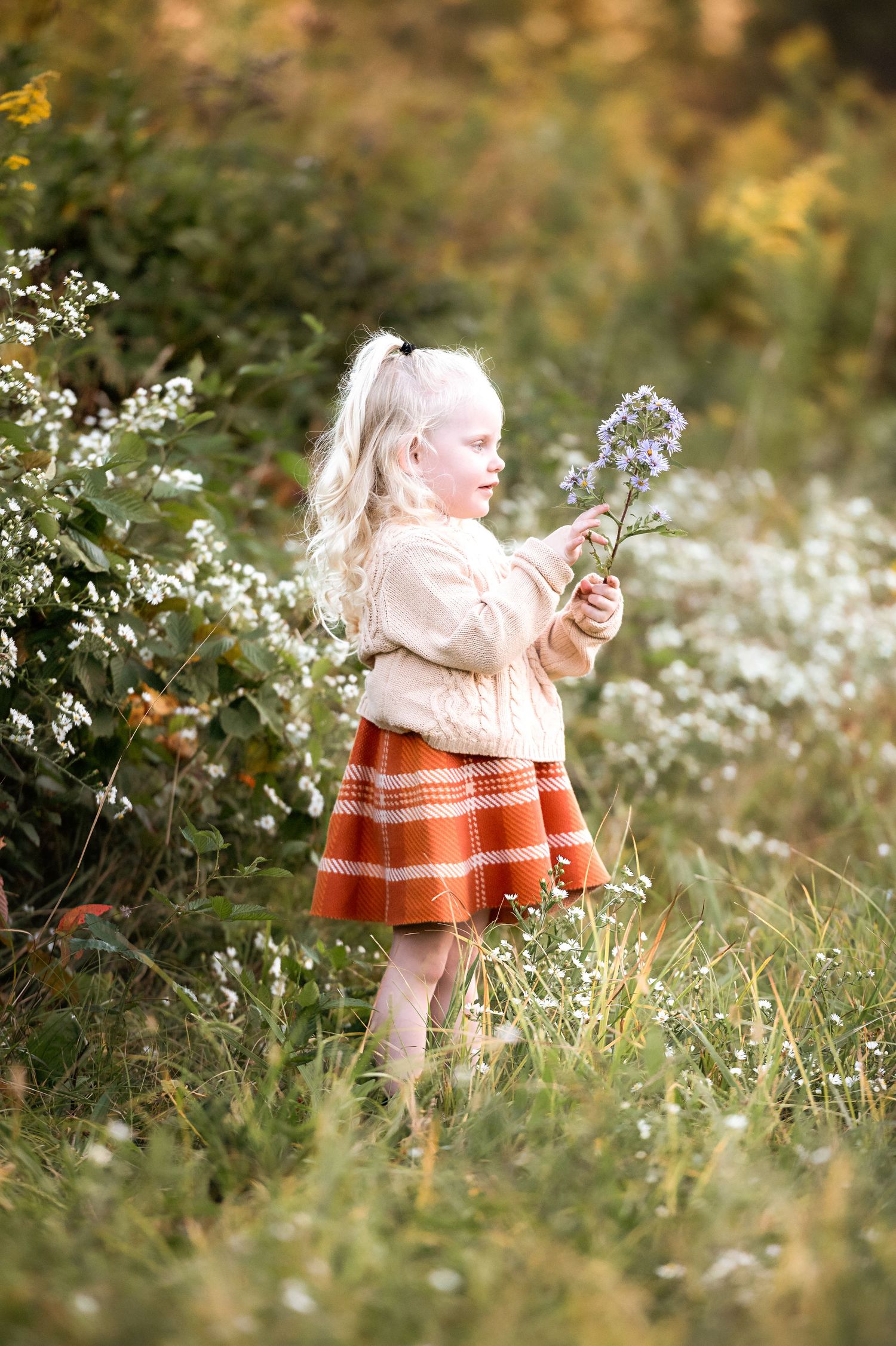 A person in an orange sweater sits among delicate white wildflowers in a dreamy outdoor setting during golden hour.