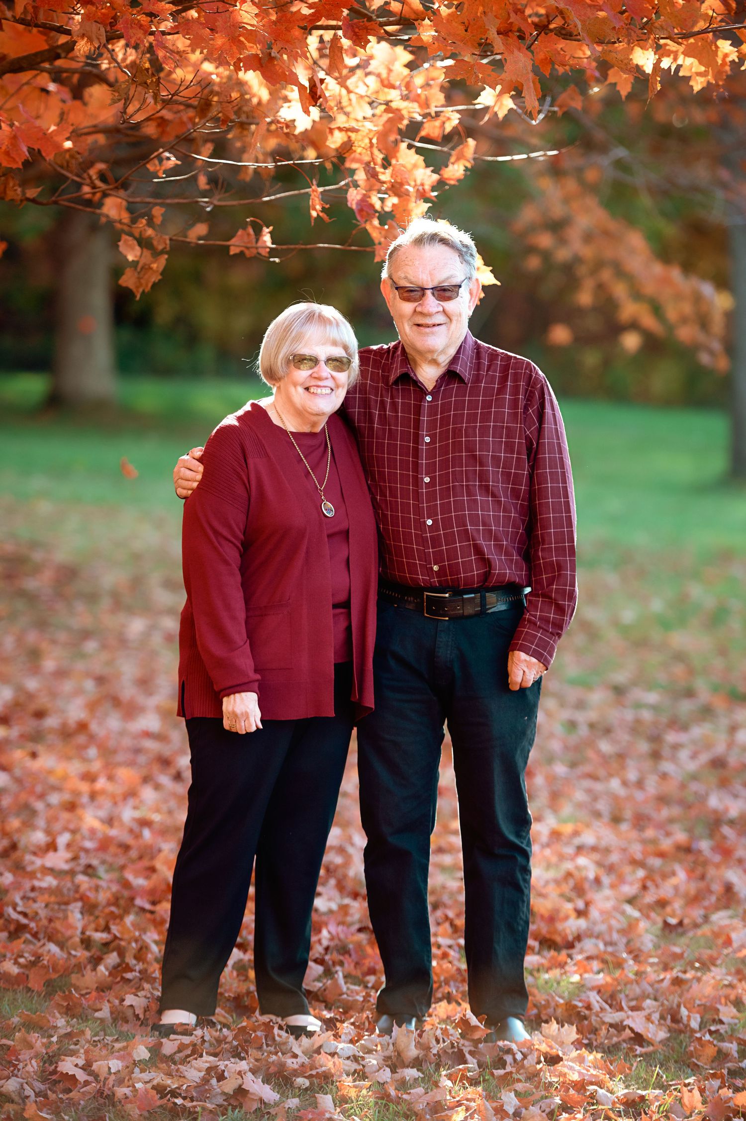 A couple stands beneath bright orange autumn leaves wearing coordinating burgundy tops.