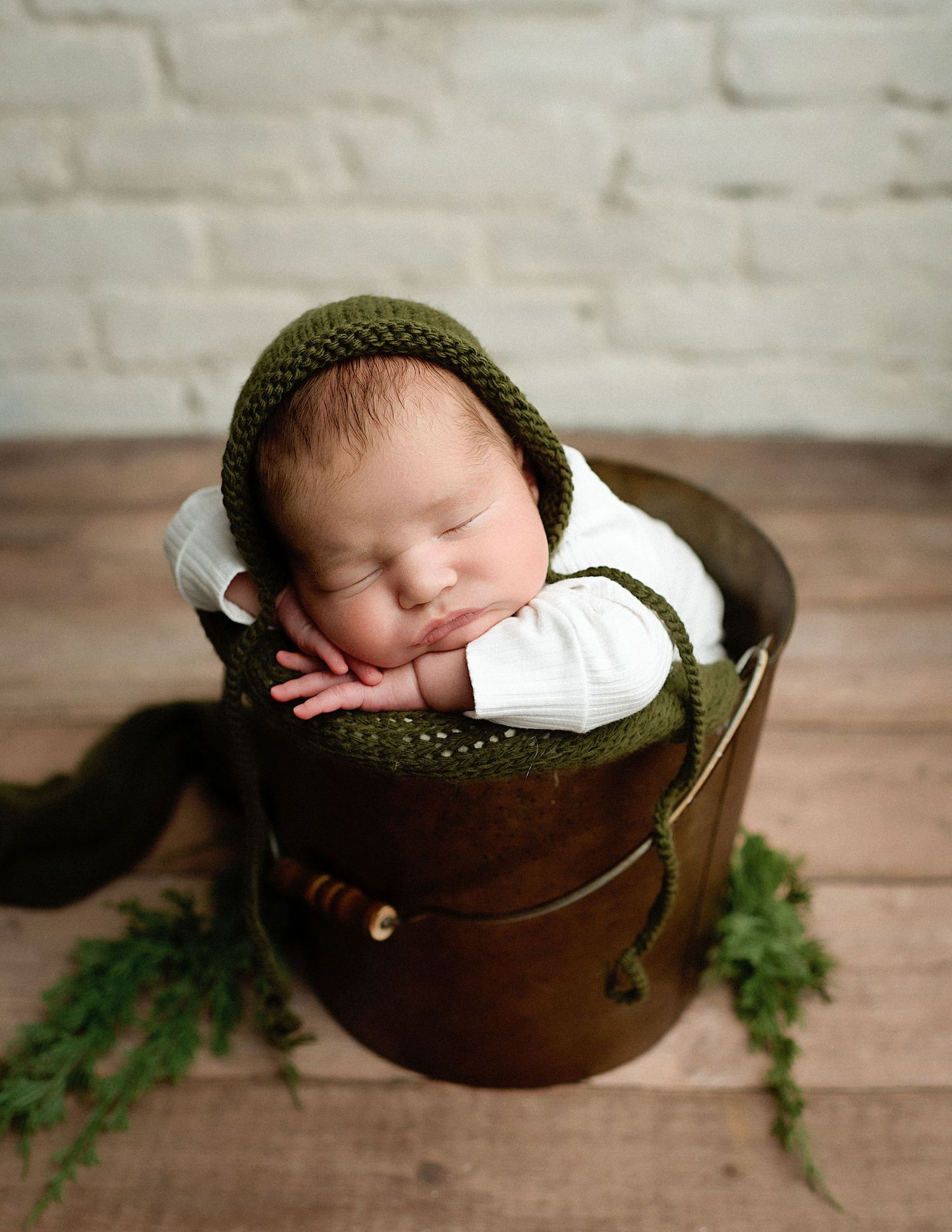 Sleeping newborn wearing green knit hat rests in antique brown leather container with greenery.