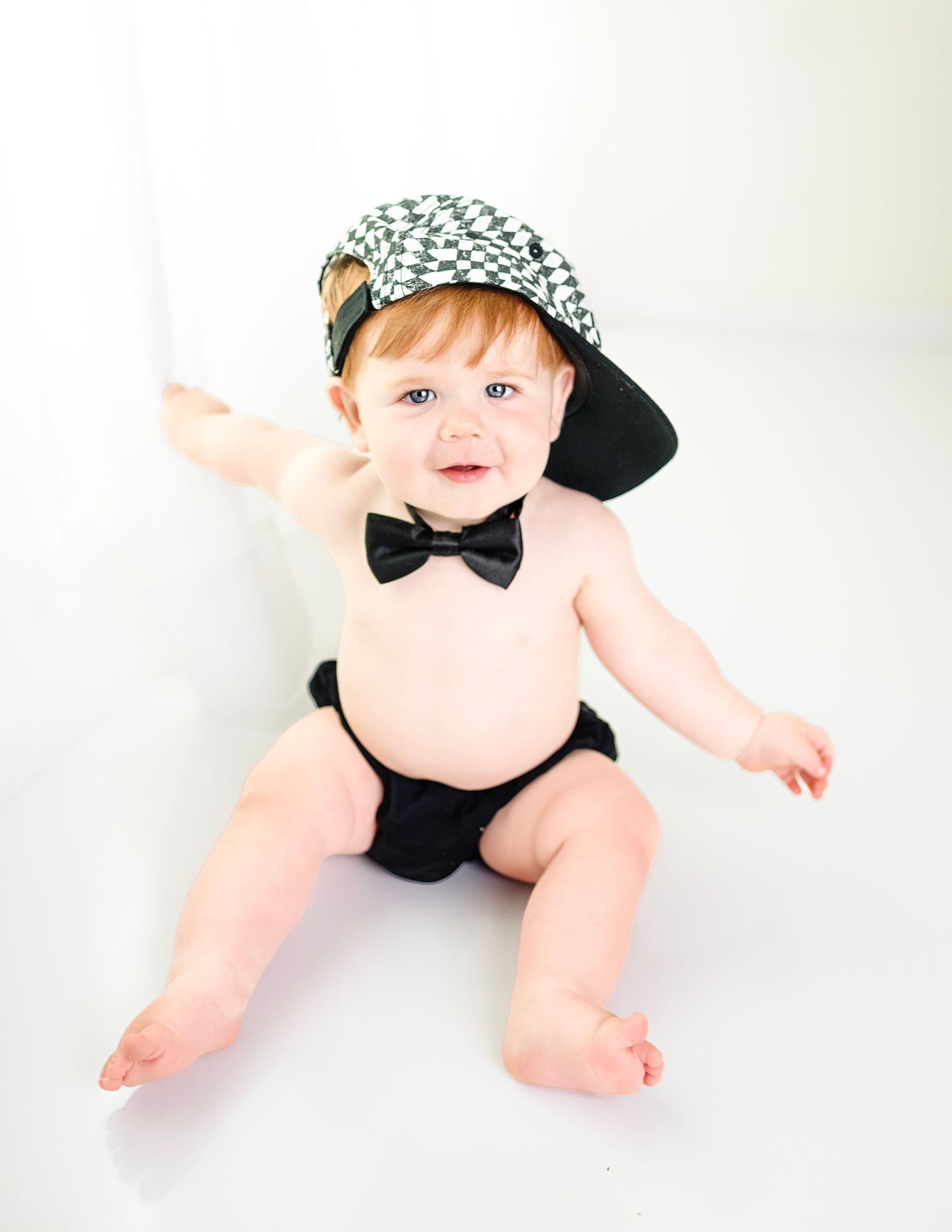Baby wearing baseball cap and bow tie poses in diaper for stylish portrait.