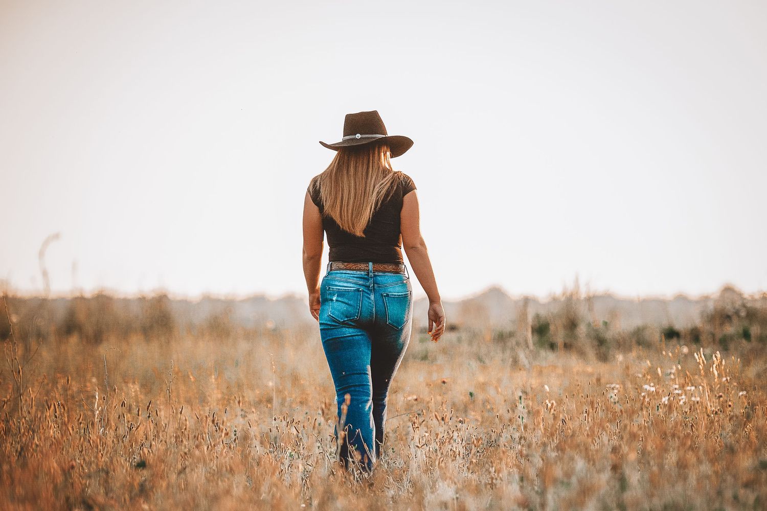 Person walking through golden field wearing wide-brimmed hat and blue jeans.