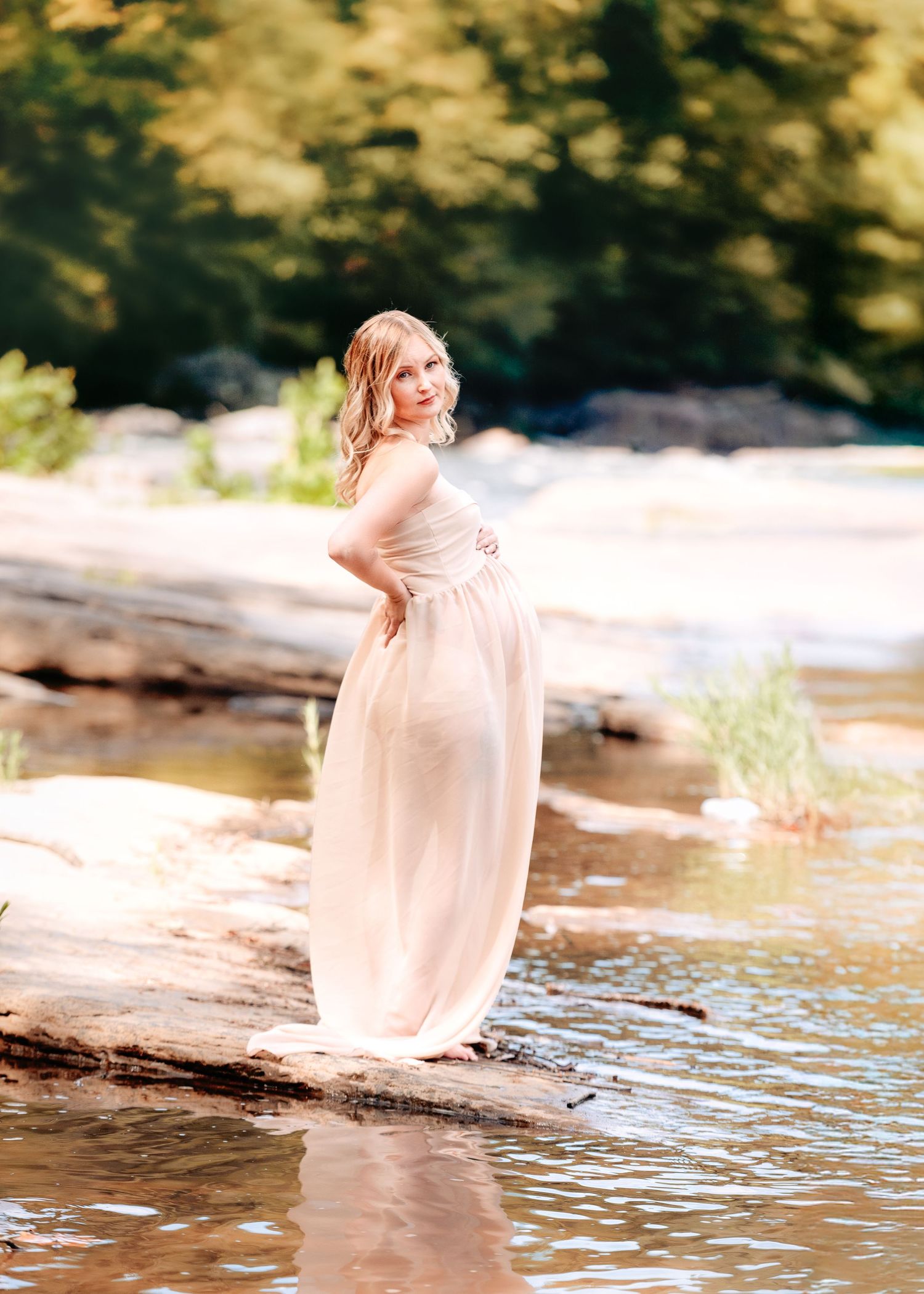 Ethereal maternity portrait by the water's edge with flowing dress and natural scenery.