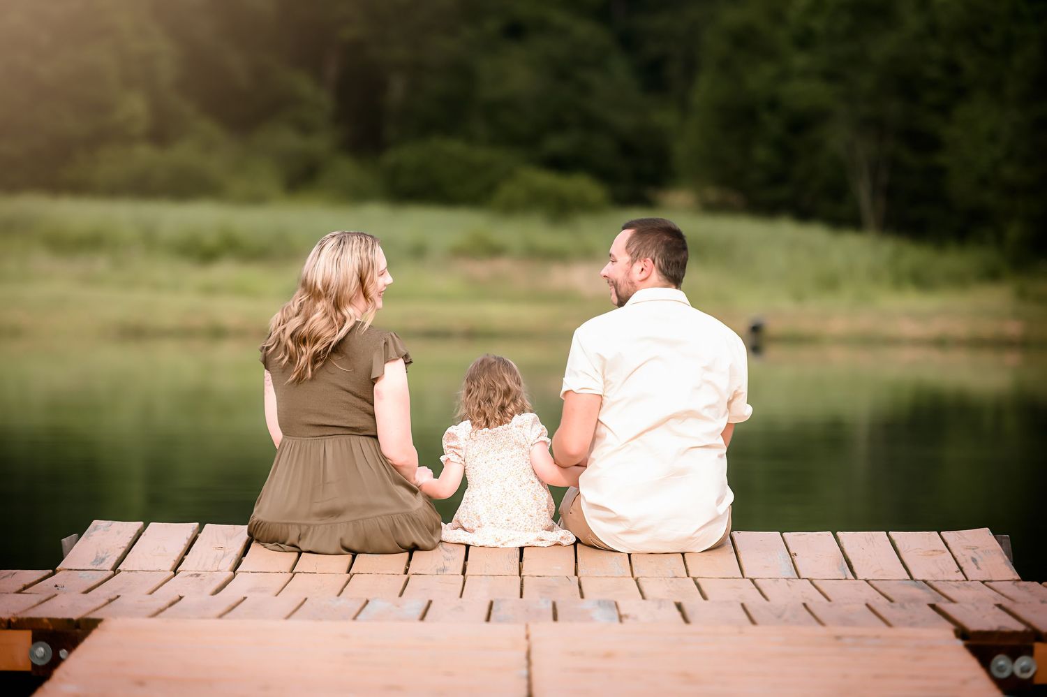A couple and child sit together on a wooden dock by a serene lake during sunset.